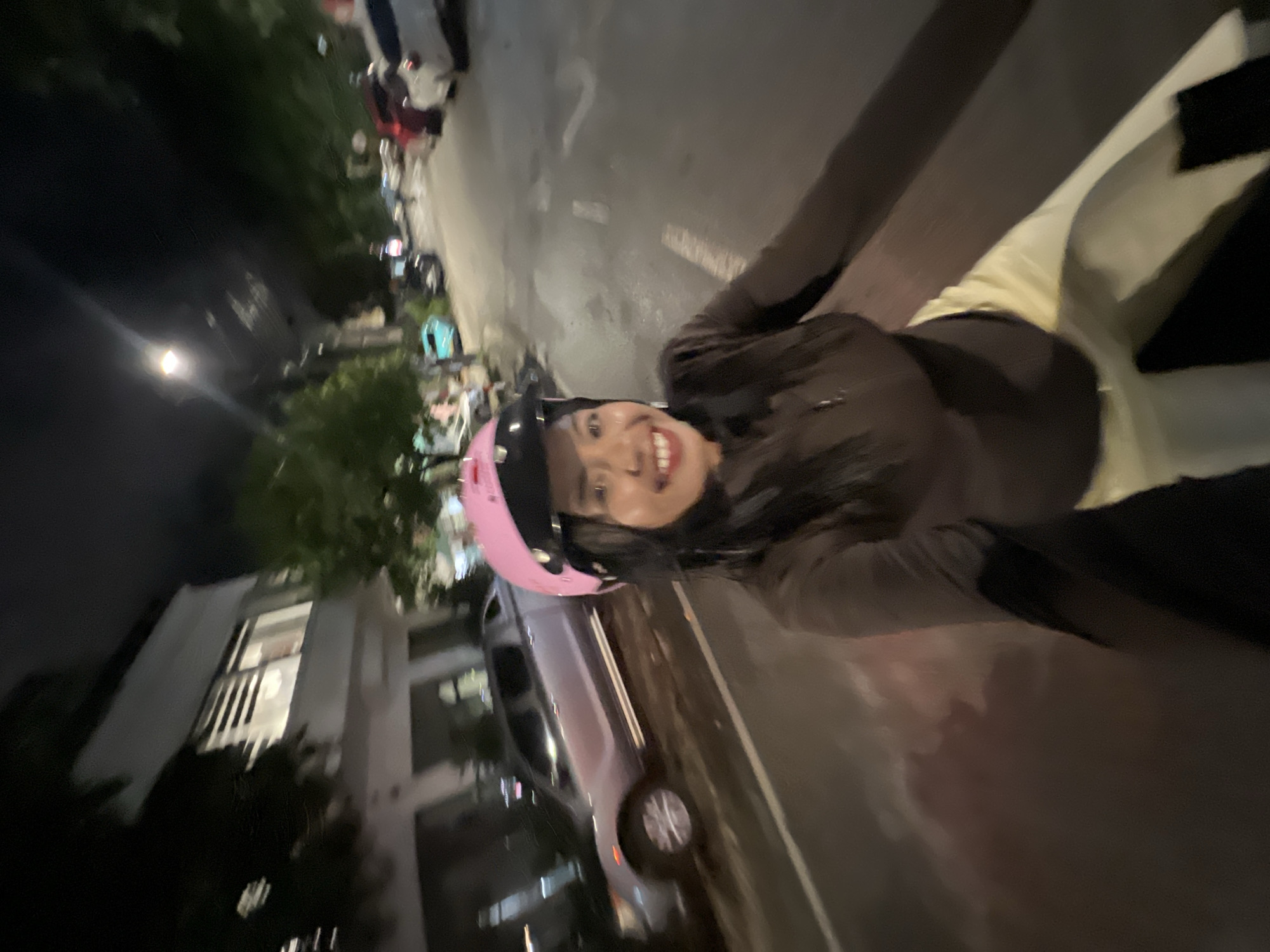 This is a first-person perspective, night time, selfie-style photo taken from a moving vehicle. A person wearing a brown zip-up jacket and light-colored pants is visible. They are wearing a pink helmet with spikes on top and have dark hair. They are smiling directly at the camera. In the background, a street is visible with parked cars, trees, and buildings. The scene appears to be in a residential or commercial area with streetlights and the glow from building windows. The location is Minh Xuân Ward, Vietnam.