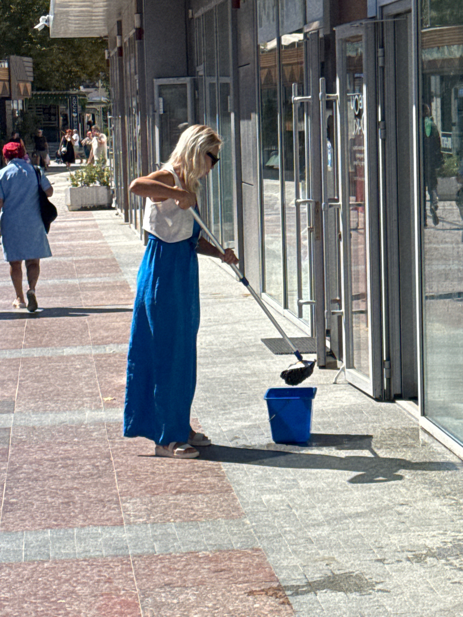 In Anapa, Russia, a woman with blonde hair, wearing a white top and a long blue dress, is mopping the paved sidewalk in front of a row of storefronts. She is holding a mop with a dark mop head, and a blue bucket sits on the ground nearby. The scene appears to be a commercial area, with glass doors and windows of shops lining the walkway. The sun is casting shadows on the ground, indicating it is daytime. Another person, wearing a blue shirt and a red hat, is walking away from the camera on the sidewalk, carrying a dark bag. Several other people are visible in the background, further down the sidewalk, walking in the same direction. The walkway is paved with a pattern of reddish and gray rectangular tiles.