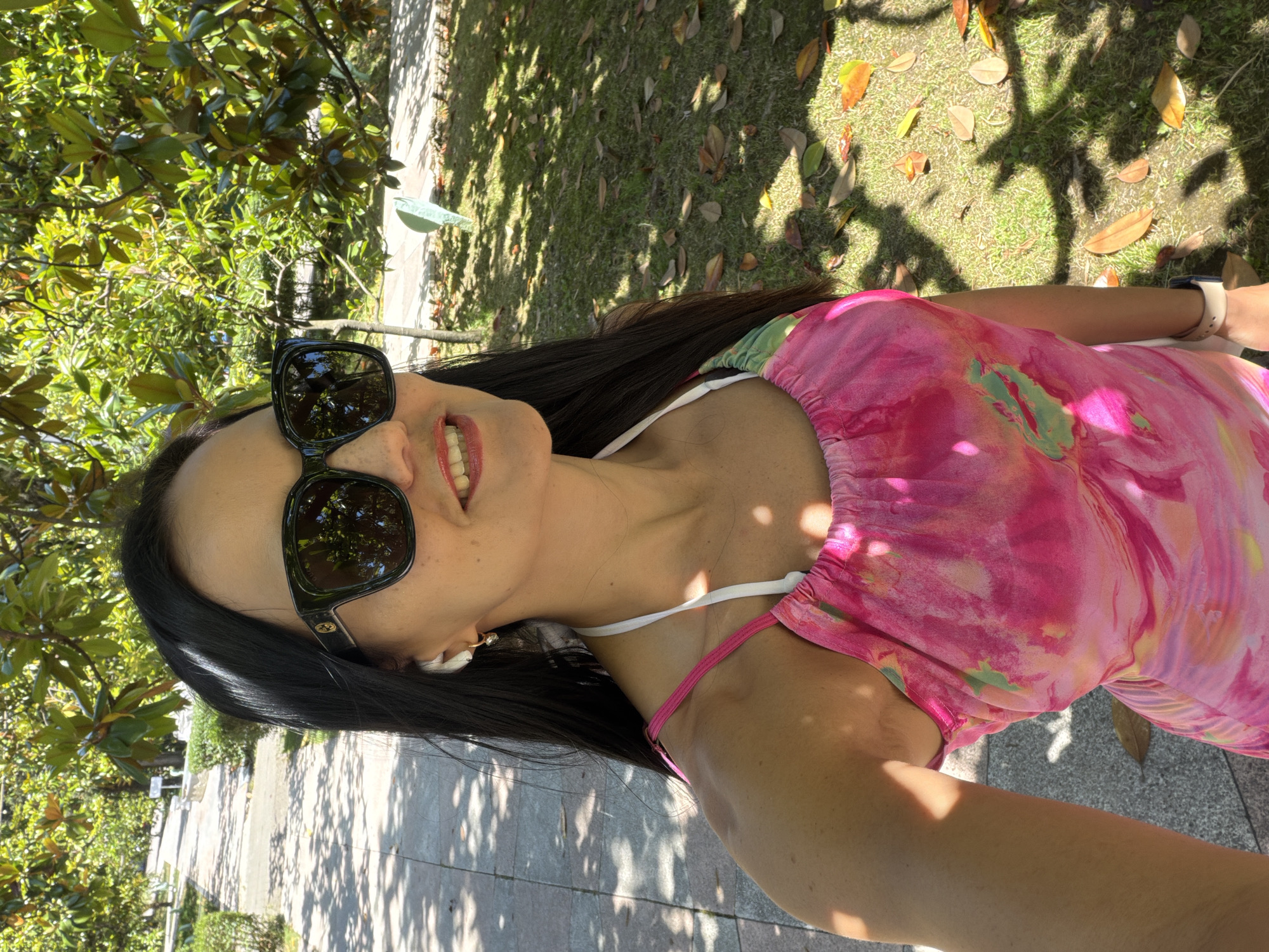 A woman with long, dark hair and sunglasses is taking a selfie outdoors. She is wearing a pink, tie-dye print dress and white earbuds. The background shows a tree-lined path with dappled sunlight on the ground. The location is a park in the city of Sochi, Russia.
