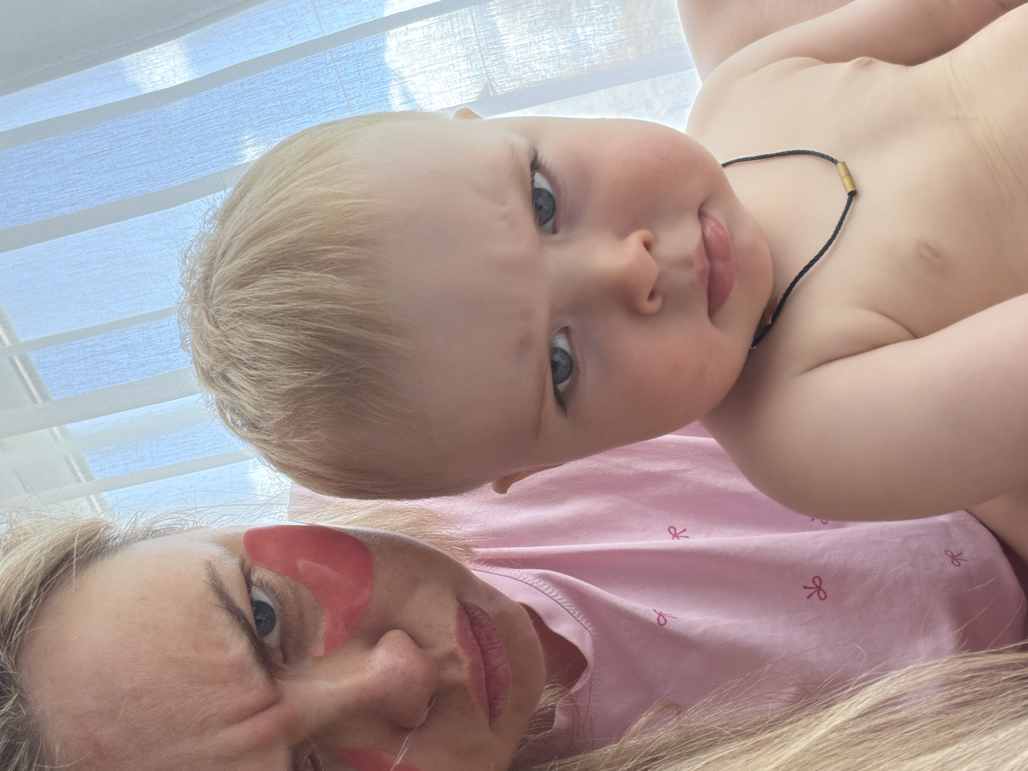 A close-up shot shows a woman and a baby. The woman is lying down, wearing pink eye patches, and a pink shirt with small bow patterns. The baby is positioned above her, looking at the camera. The baby has fair skin and blonde hair and is wearing a dark necklace. The background consists of a window with blinds. The location is indiscernible and cannot be reliably determined to be in Votkinsk, Russia based on the image content.