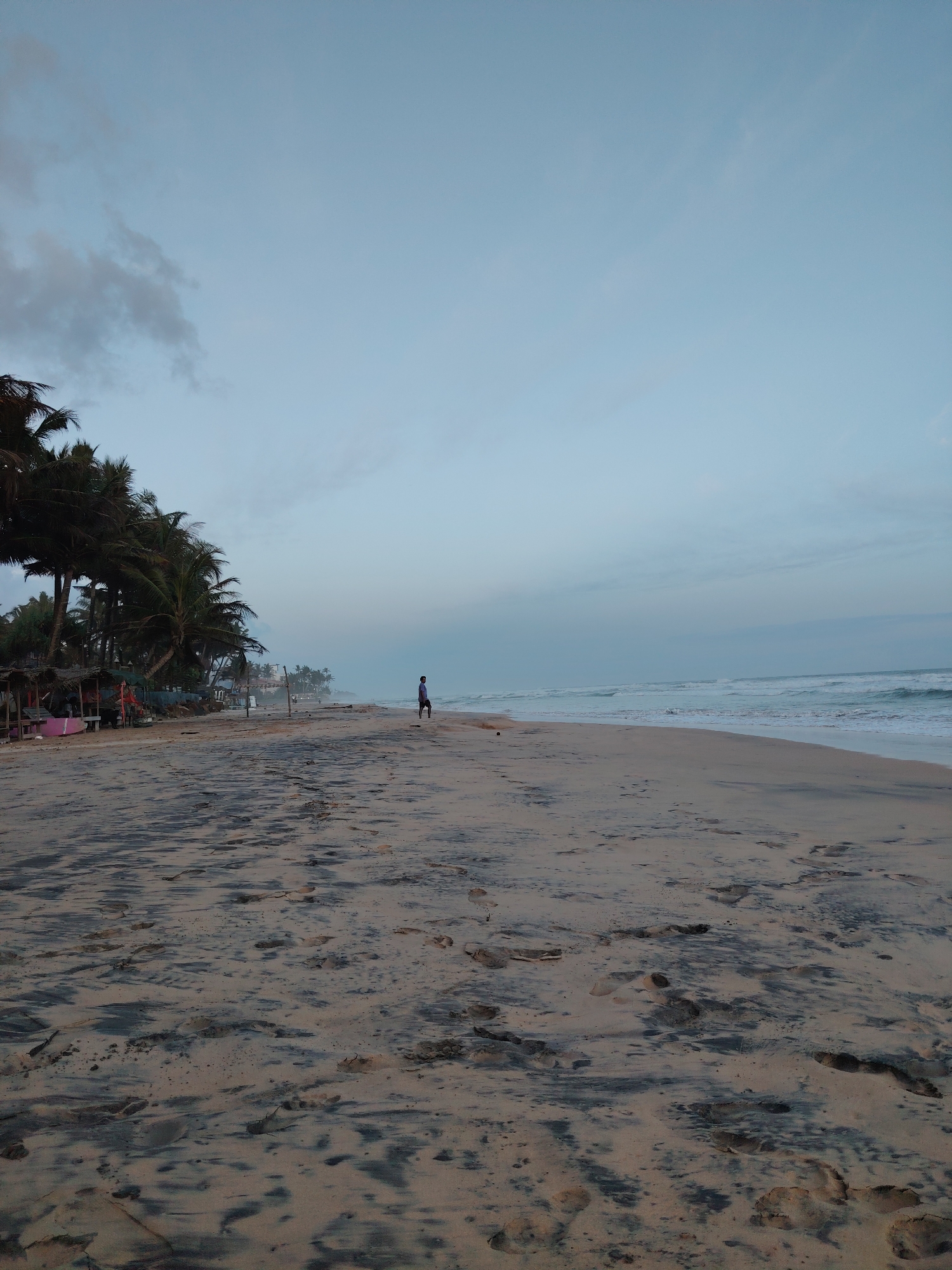 A lone individual stands on a sandy beach, facing the ocean. The sky is overcast, and the waves are gently rolling in. The beach is littered with footprints, and palm trees line the shore in the distance. There are also some small huts or shelters visible, suggesting a tropical location.