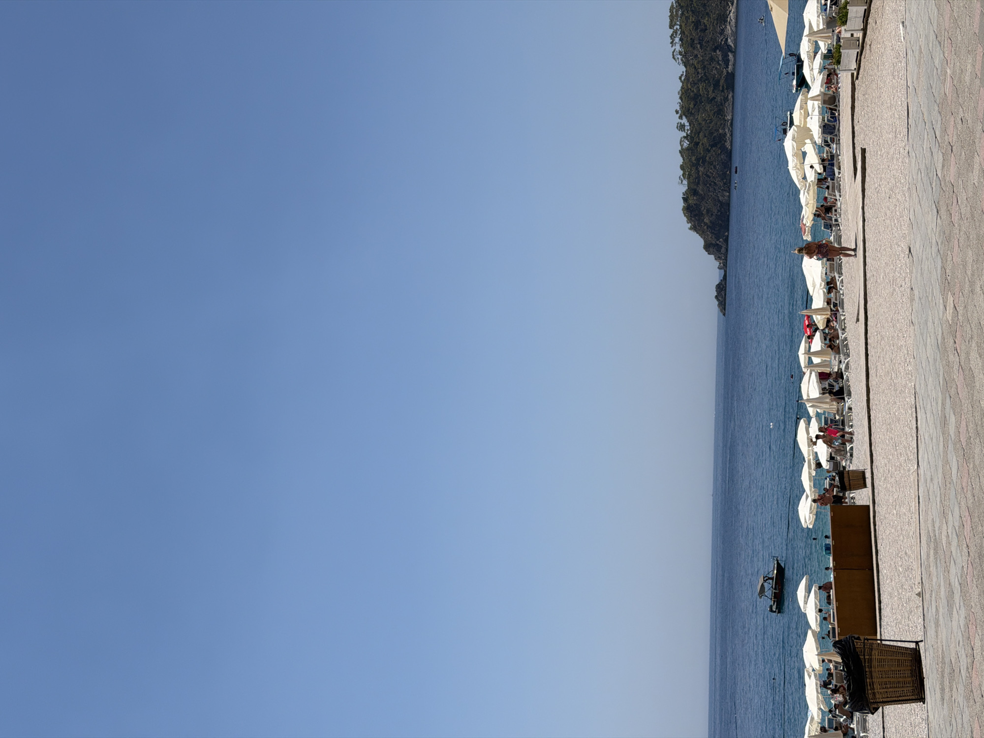 A bright, sunny day at a beach in Fethiye, Turkey is depicted. The clear blue sky dominates the upper half of the image. The sea is a deep blue, with a small boat visible in the distance near a rocky, tree-covered peninsula. The foreground is a beach with rows of white umbrellas providing shade for lounge chairs. Many people are seen relaxing on the beach, some in swimwear, while others are standing or walking. A woman in a patterned swimsuit stands near the center of the image, facing away from the camera. Two large wicker trash cans are present on the left side of the foreground. The beach area is paved with light-colored stones, and beyond that is a tiled walkway. The overall atmosphere is one of a popular tourist destination on a warm day.