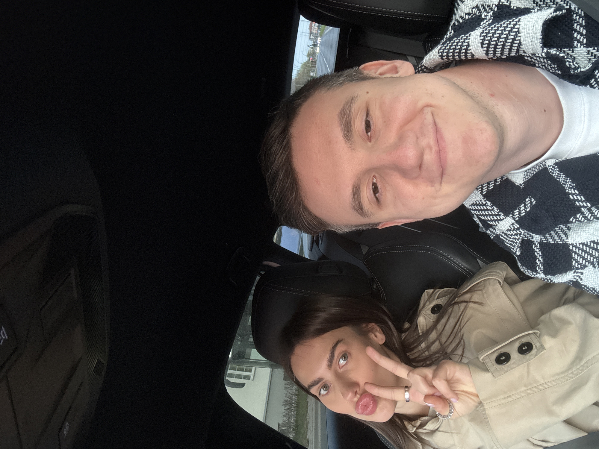 Two individuals are in the front seats of a car. The man is wearing a white t-shirt and a black-and-white checkered jacket. The woman is wearing a beige trench coat and making a peace sign with her hand. The interior of the car is black. Outside the car, a building is visible. The picture was not taken in Warsaw, Poland.