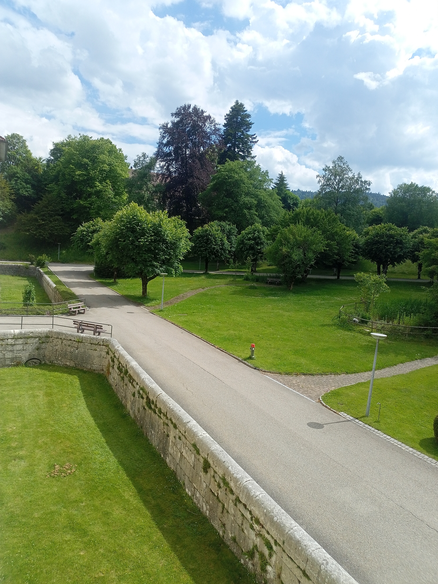 This media file captures an outdoor scene on a partly cloudy day. The primary subjects are the lush green landscape, a paved pathway, and a stone wall. The pathway curves through a well-maintained park with green grass and scattered trees. On the pathway, there are two wooden benches visible, suggesting a place for rest and contemplation. A fire hydrant is also present near the edge of the path. The stone wall, constructed with large, rough-hewn blocks, runs along the foreground and appears to be an old fortification or retaining wall. Beyond the immediate park area, a dense forest of trees can be seen, with some taller, darker trees standing out. The sky is a mix of blue sky and fluffy white clouds, indicating fair weather. The lighting suggests it is daytime, likely late morning or early afternoon, given the shadows cast by the trees and the light intensity. There is no text visible in the image. The overall impression is one of a serene and picturesque park setting, possibly in a historical or natural area.