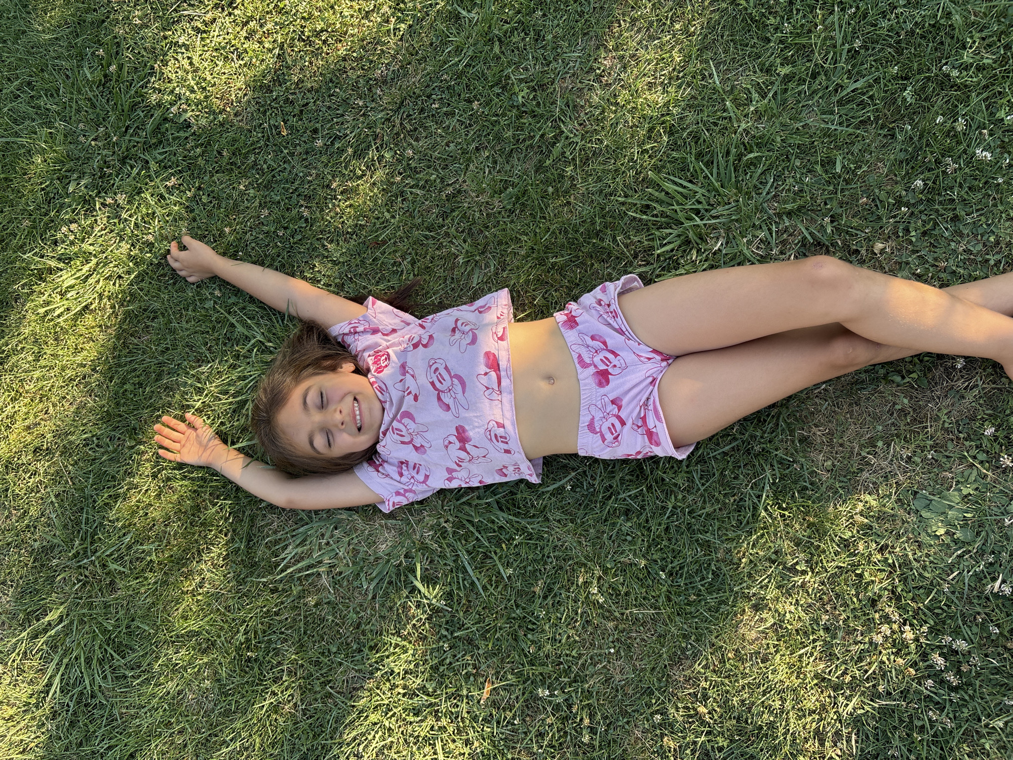 A young girl lies on her back in a grassy field, her arms raised above her head. She is wearing a pink t-shirt and shorts with a cartoon character print. Her eyes are closed, and she appears to be smiling. The grass is green and lush, with some patches of clover and small white flowers. Sunlight filters through the trees, casting dappled shadows on the ground. The scene suggests a moment of relaxation and enjoyment in a natural outdoor setting. The context provided indicates this scene is in Oreshak, Bulgaria.