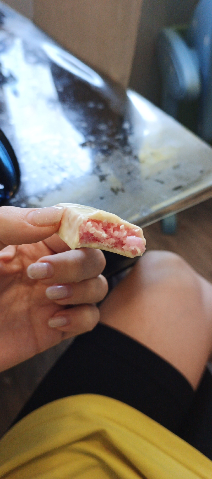 A person is holding a partially eaten white chocolate-covered snack with a pink filling. The person has light-colored fingernails and is wearing a yellow shirt and black shorts. The background includes a shiny, metallic surface and a glimpse of a light blue object. The image's location in Chaikovsky, Russia, cannot be determined from the visual information alone.
