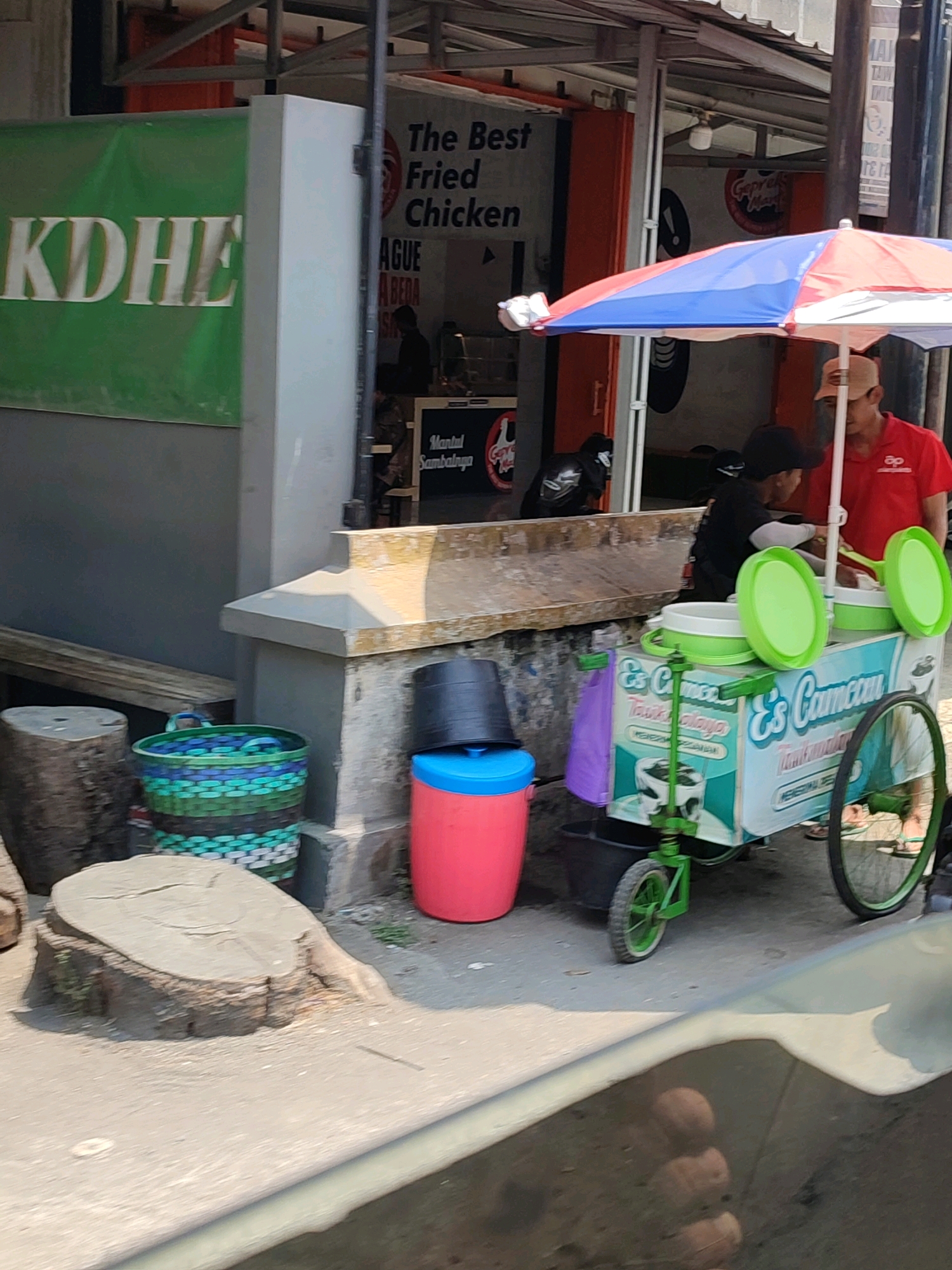 The image depicts a street scene in Secang, Indonesia, with a food stall prominently featured. In the background, a sign reads "The Best Fried Chicken." A man in a red shirt and a tan cap is working at a colorful ice cream cart, which has "Es Camcau" written on its side. Another person, wearing a black shirt and cap, is also at the cart. Two large buckets, one black and one pink, are near the cart. In the foreground, a woven basket with green and blue patterns sits on the ground. To the left, a tree stump is visible. The ground is paved, and a portion of a vehicle's windshield is in the lower part of the frame, reflecting the surroundings.