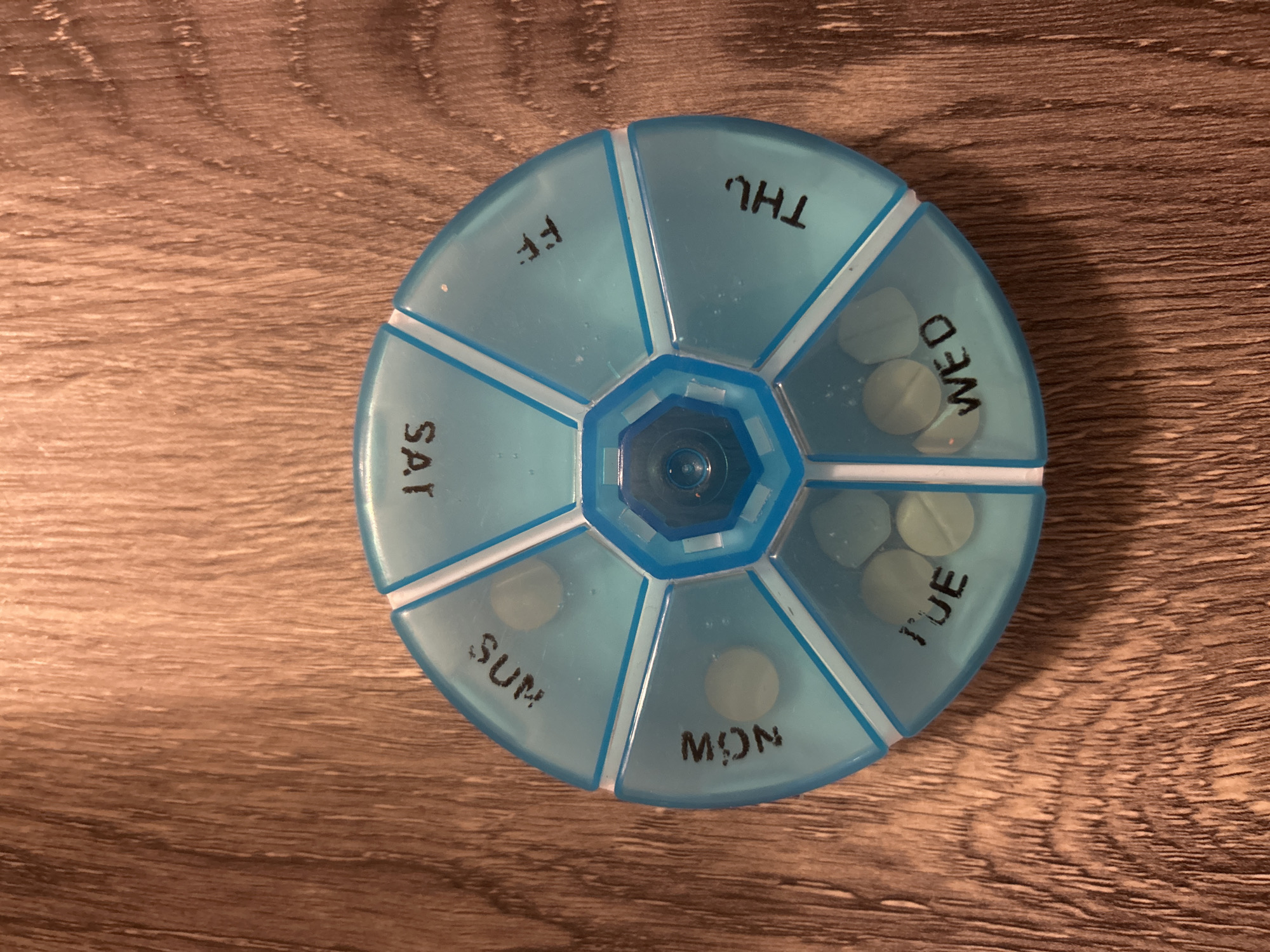 A blue, circular pill organizer rests on a brown wooden surface. The organizer has seven compartments labeled with the days of the week: 'FF', 'THU', 'WED', 'TUE', 'MON', 'SUN', and 'SAT'. Several compartments contain round, light-colored pills. The center of the organizer has a blue, octagonal hub. The surface appears to be a table or countertop, featuring a wood grain pattern. The setting is indoors with soft lighting. The location is impossible to know from just looking at this image.