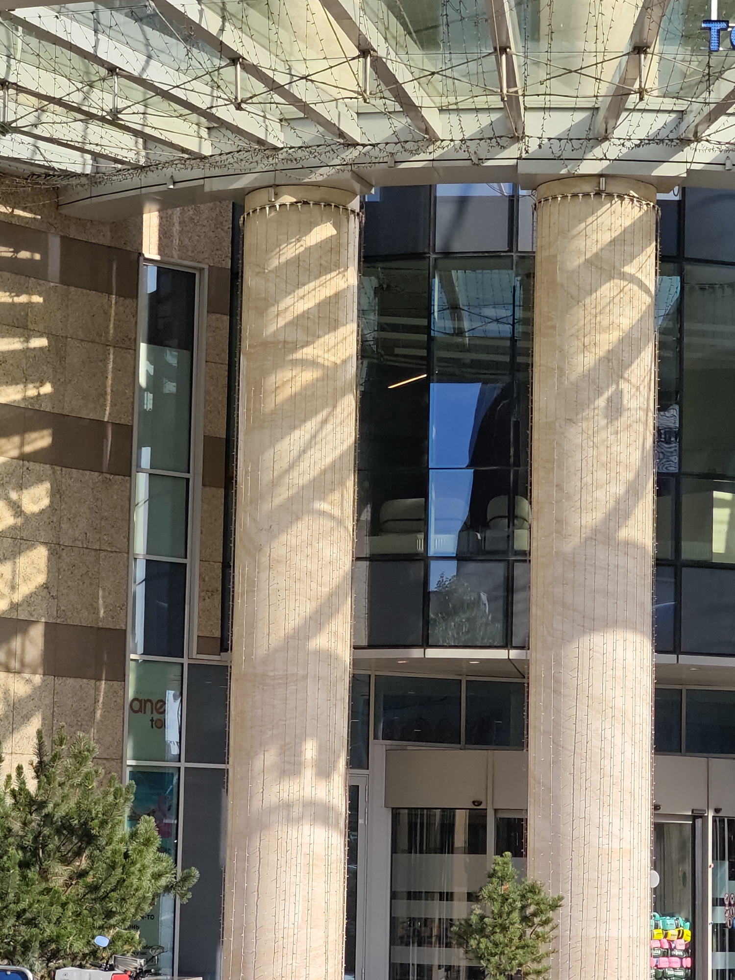 The image shows the facade of a modern building, likely a commercial or office building, with large glass windows and two large cylindrical columns made of light-colored stone. The columns are adorned with strings of small lights. The building's exterior wall is a mix of beige stone tiles and darker brown horizontal accents. Sunlight casts diagonal shadows from the roof structure onto the columns and the building's facade. In the foreground, on the left side, a portion of a green coniferous tree is visible, and at the very bottom left, a part of a motorcycle with a blue mirror is partially in frame. Through the glass windows, reflections of the sky and surrounding environment are visible, as well as some interior elements like furniture and signage. A sign on one of the lower windows appears to read "one to". The architecture suggests an urban setting. The text "TO" in blue is visible at the top right, suggesting this is a building in Moscow, Russia.