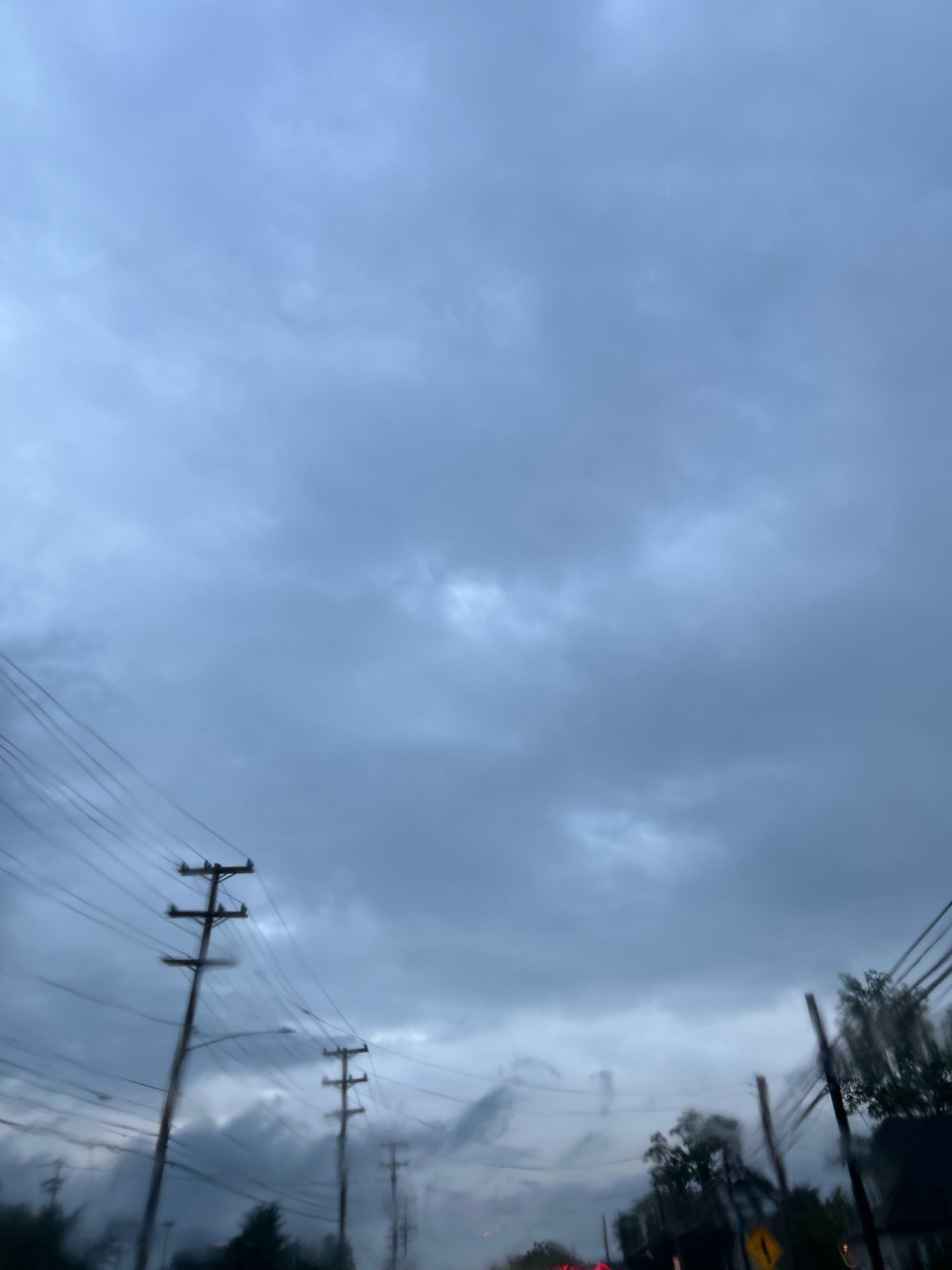 A low-angle, slightly blurred photograph captures a view of utility poles and power lines against a heavily overcast, grey sky. The scene appears to be from a moving vehicle, as indicated by the motion blur on the foreground elements and the perspective looking upwards. Several utility poles, characterized by their wooden structure and crossbars holding numerous wires, are visible. The sky is filled with dense, dark clouds, suggesting a gloomy or impending weather event. Silhouetted trees and glimpses of other structures, possibly houses or businesses, are visible at the bottom of the frame, further suggesting an urban or suburban environment. The overall impression is one of a typical cloudy day in an area with overhead power infrastructure.