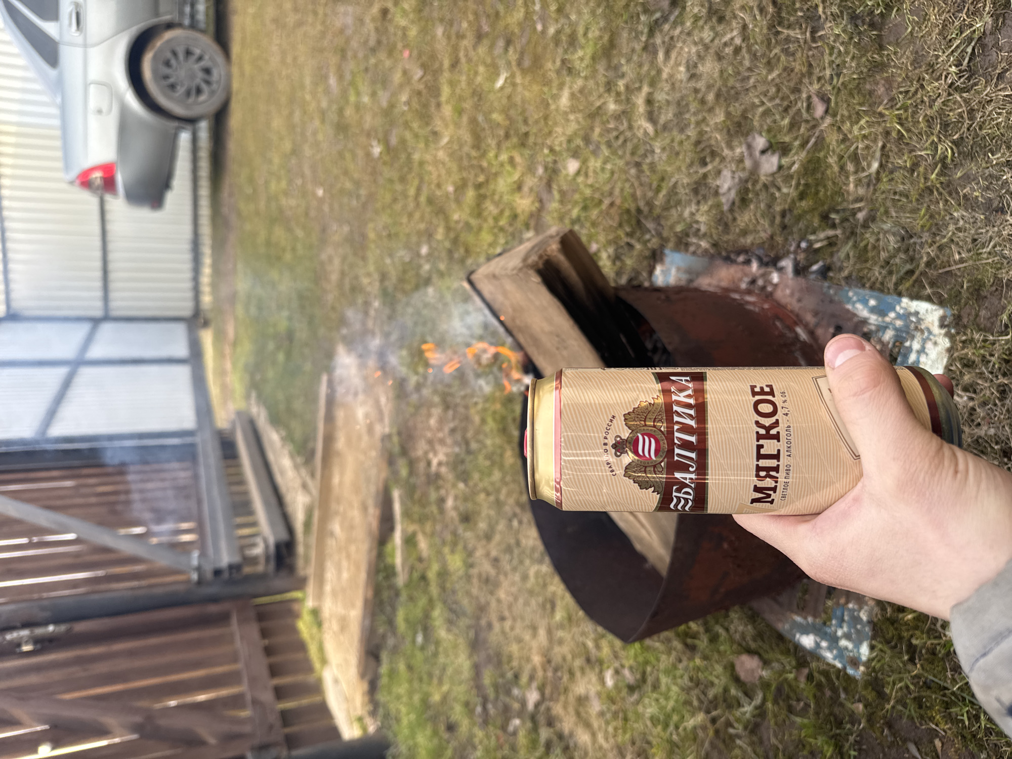 The image, taken in Бородулино, Russia, presents an outdoor scene centered around a hand holding a beverage can above a small fire. The photograph is oriented sideways, with the right side of the image being the top.
The main subject is a human hand, likely a right hand, gripping a gold-colored aluminum can. The can is clearly branded with "БАЛТИКА" (Baltika) and features the word "МЯГКОЕ" (Myagkoye, meaning Soft) in a prominent font. Additional text on the can identifies it as "СВЕТЛОЕ ПИВО" (Light Beer) with an "АЛКОГОЛЬ - 4,7% об." (Alcohol - 4.7% vol.) content, and states "СВАРЕНО в РОССИИ" (Brewed in Russia).
Directly below the can, a small fire burns within what appears to be a rusty, cut-off metal barrel or container. Logs or pieces of wood are visible inside the barrel, feeding the flames, which are bright orange and red, emitting a plume of light smoke.
The background consists of a grassy ground, indicating an outdoor, possibly rural or suburban, setting. In the upper left portion of the image (when viewed upright), the rear section of a silver car is visible, parked alongside a corrugated metal fence or the wall of a shed. Another wooden structure is partially visible further to the left. The natural lighting suggests it is daytime with clear weather. The scene portrays a casual moment, possibly during an outdoor gathering or a relaxed activity involving a small bonfire.