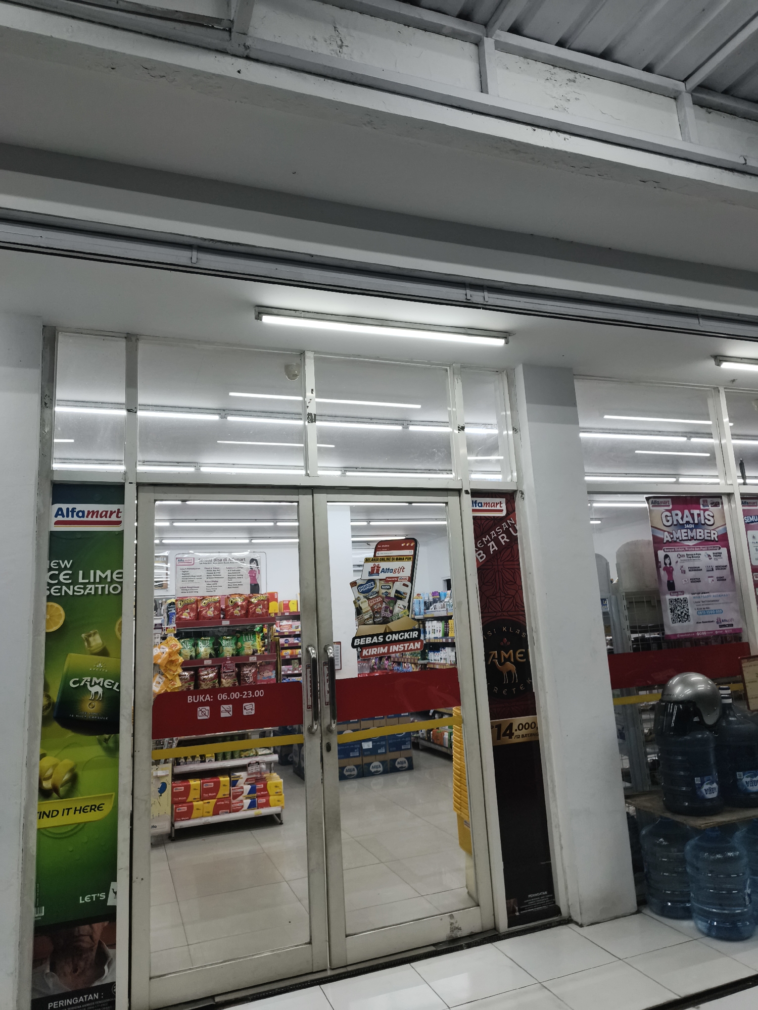 The image shows the exterior of an Alfamart convenience store in Sidorejo, Indonesia. The store has large glass doors and windows, revealing shelves stocked with various products. A green advertisement for Camel cigarettes is displayed on the left side of the entrance, featuring a depiction of lemons and ice. A red banner across the doors indicates the store's operating hours, from 06:00 to 23:00. Inside, a variety of snacks and drinks are visible on the shelves. To the right of the entrance, a poster advertises "GRATIS A-MEMBER" (Free A-Member). Further to the right, stacks of large water bottles are arranged near the entrance, one topped with a helmet. The floor appears to be tiled in a light color. The ceiling is made of metal panels with fluorescent lighting fixtures. The overall impression is that of a typical Indonesian convenience store operating during daylight hours.