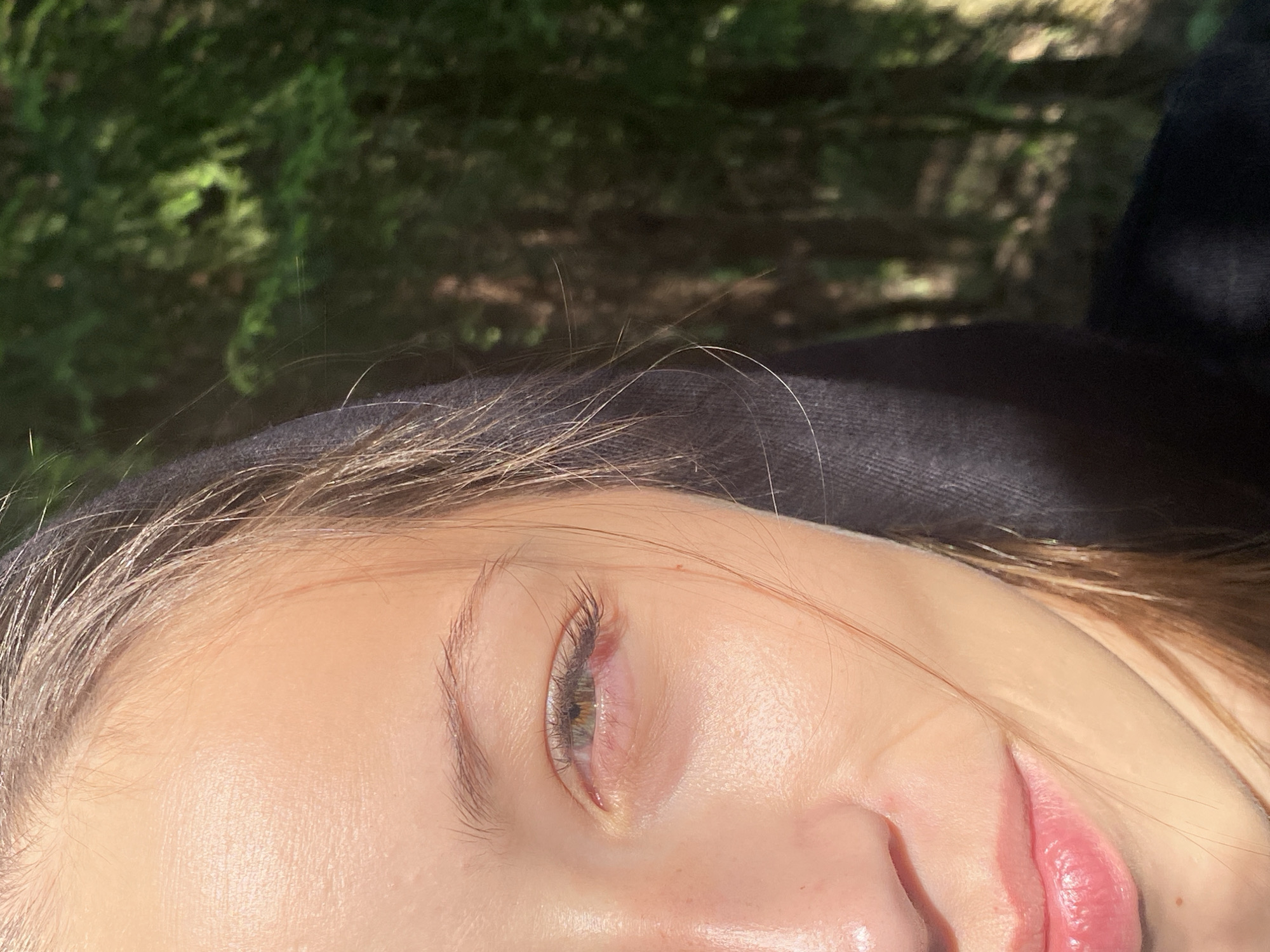 This close-up image presents a tilted, upward-looking perspective of the upper right side of a person's face, illuminated by strong, direct sunlight. The subject has fair skin, highlighted by the sun, and light brown or blonde hair with fine, light strands visible across the forehead and against a dark fabric.
The person's right eye is partially closed, showing a hazel or light green/brown iris and long, dark eyelashes. The skin around the eye has subtle folds, suggesting a relaxed or contemplative expression. A portion of the nose and the top of naturally pink lips are also visible.
The person appears to be wearing a dark, possibly black, casual garment, such as a hoodie or jacket, which frames the lower part of the face and hair. The background is a heavily blurred expanse of green and brown foliage, indicating an outdoor, natural setting like a forest or garden. The bright sun suggests it is daytime. No text is visible. The outdoor context aligns with the known natural beauty of Komarovo, Russia.