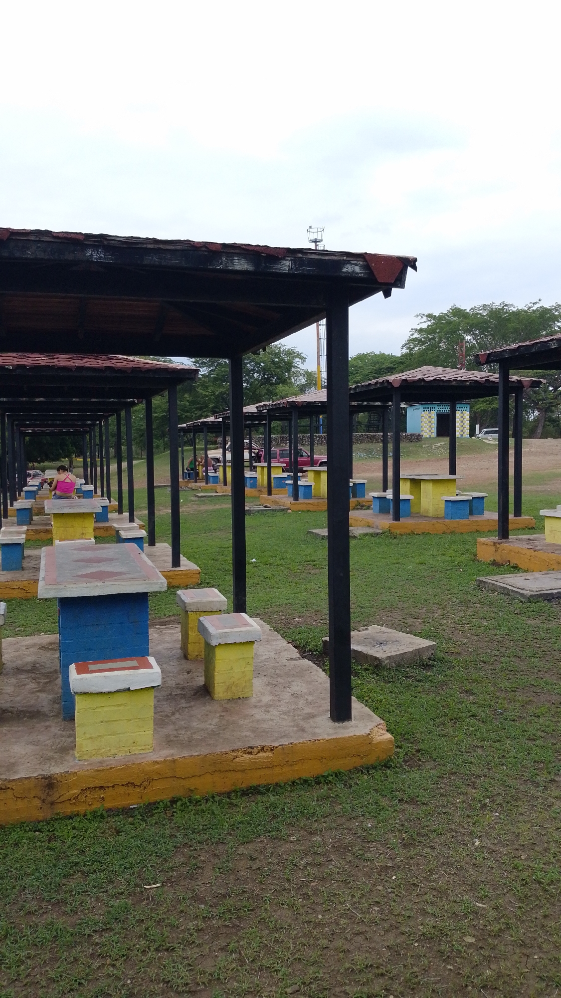 The image depicts a park setting in Tinaquillo, Venezuela, featuring rows of picnic tables under covered structures. The tables are constructed of concrete, painted with a combination of yellow and blue colors. The coverings are supported by black metal beams. The ground is a mix of grass and bare earth. In the distance, there are trees and a light blue building. Several people are visible occupying some of the picnic tables. The sky is overcast.