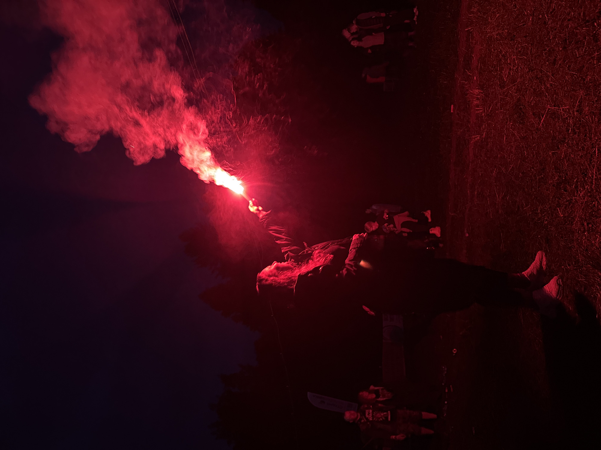 The image depicts an outdoor nighttime scene in Большая Кивара, Russia. A central figure, appearing to be a person with long hair, is positioned in the middle right of the frame, holding a luminous red flare aloft with their right hand. The flare emits a dense, bright red smoke cloud that billows upwards and to the right, intensely illuminating the person's profile and the immediate surrounding area with a deep red light. The base of the flare exhibits an almost white-hot core.
Several other individuals are present in the background, primarily silhouetted or dimly lit by the red flare. To the far left, a person wearing a dark top with a light-colored graphic design and dark shorts is partially visible. Further back and to the left, a light-colored banner or flag with some indiscernible blue graphics is erected. In the mid-ground to the right, a small group of people stands, their forms largely obscured by shadow. Faint overhead lines or cables are visible stretching across the upper right portion of the image, passing through the red smoke.
The ground appears to be an uneven, possibly grassy or dirt surface, with scattered faint details suggesting debris. Dark tree lines form a silhouette against the slightly lighter, dark blue-purple night sky in the background, particularly on the left side of the frame. The overall environment is characterized by very low ambient light, with the red flare serving as the primary light source.