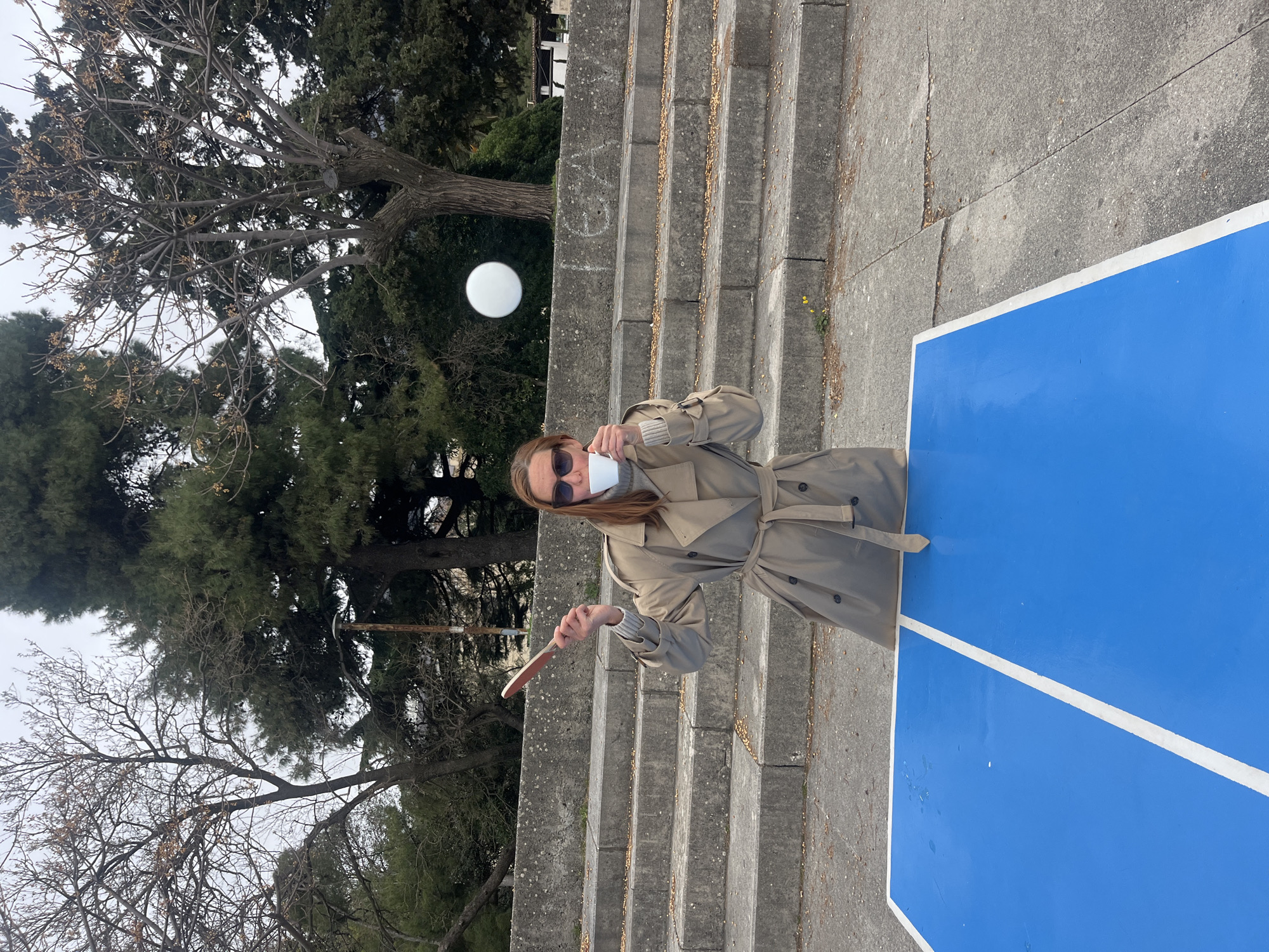 A person stands on a set of concrete stairs next to a blue-painted surface. The person is wearing a beige trench coat and sunglasses, and they are holding a white cup to their mouth with their right hand and a wooden utensil, possibly a knife or spatula, in their left hand. The background includes trees and a white sphere-shaped light fixture. The location may be Budva, Montenegro.