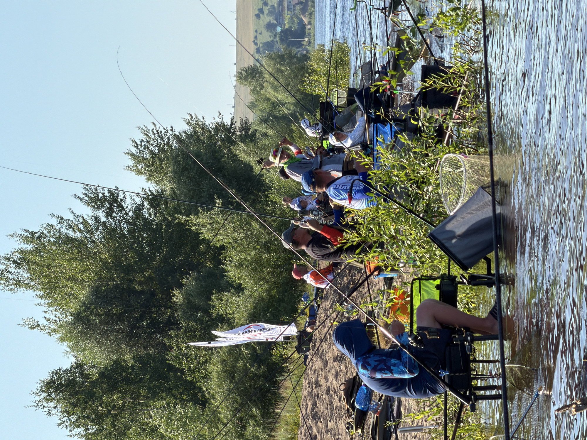A group of people are fishing on the bank of a river. The scene is set in Tarasovo, Russia, with lush green trees and a clear blue sky in the background. Several fishing rods are set up, with people sitting on chairs or stands by the water. Some individuals are actively engaged in fishing, while others appear to be waiting. The river is calm, with ripples on the surface. Various fishing equipment, such as nets and buckets, are visible near the anglers. The overall atmosphere suggests a relaxed outdoor activity.