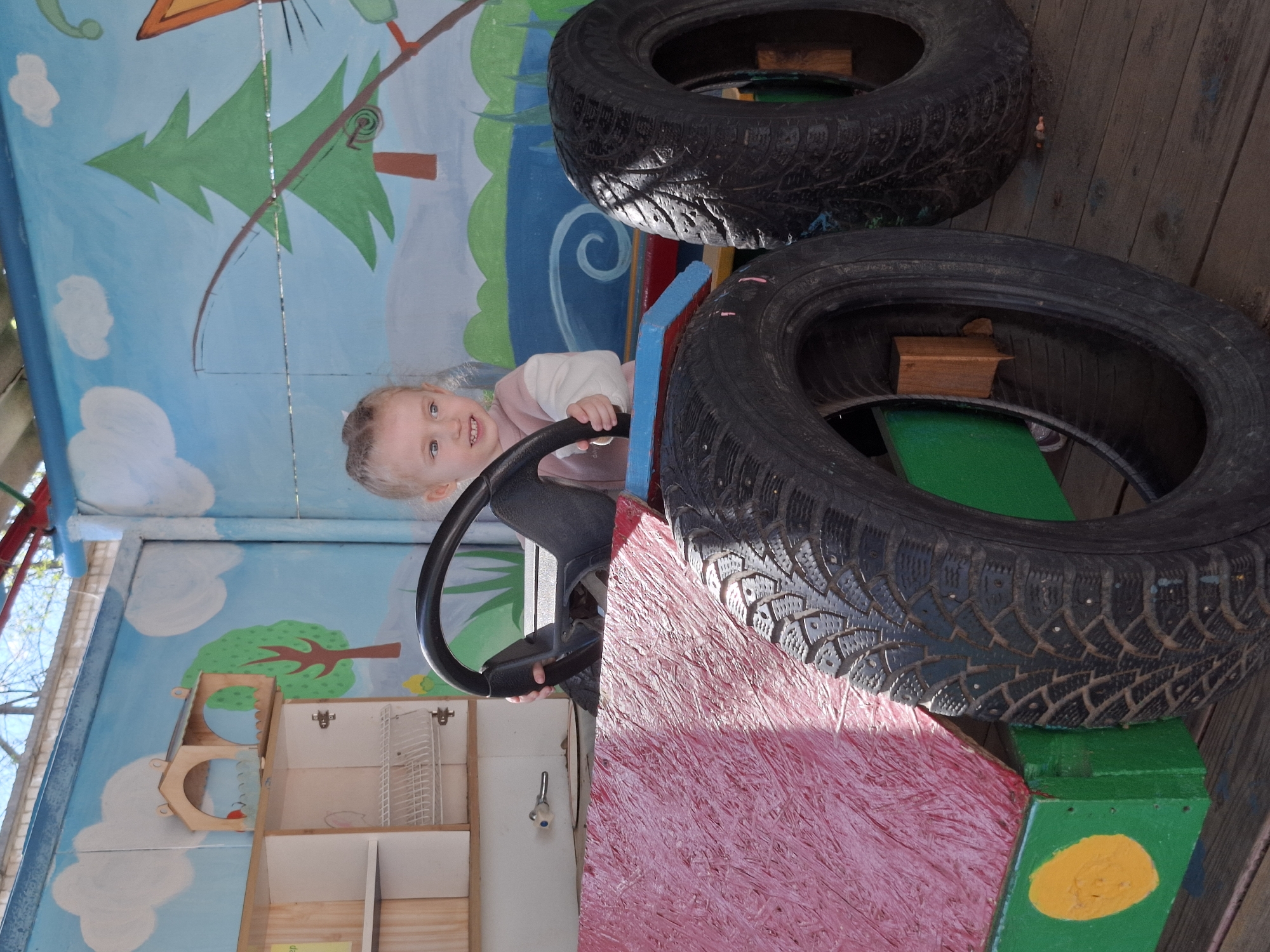 A young child sits in a makeshift, painted play structure constructed from tires in what appears to be an outdoor play area. The play structure, intended to resemble a vehicle, is painted in shades of pink, green, and blue and features a steering wheel. The background consists of a painted mural depicting trees, clouds, and a blue sky. The photo was taken in the city of Chaikovsky, Russia.