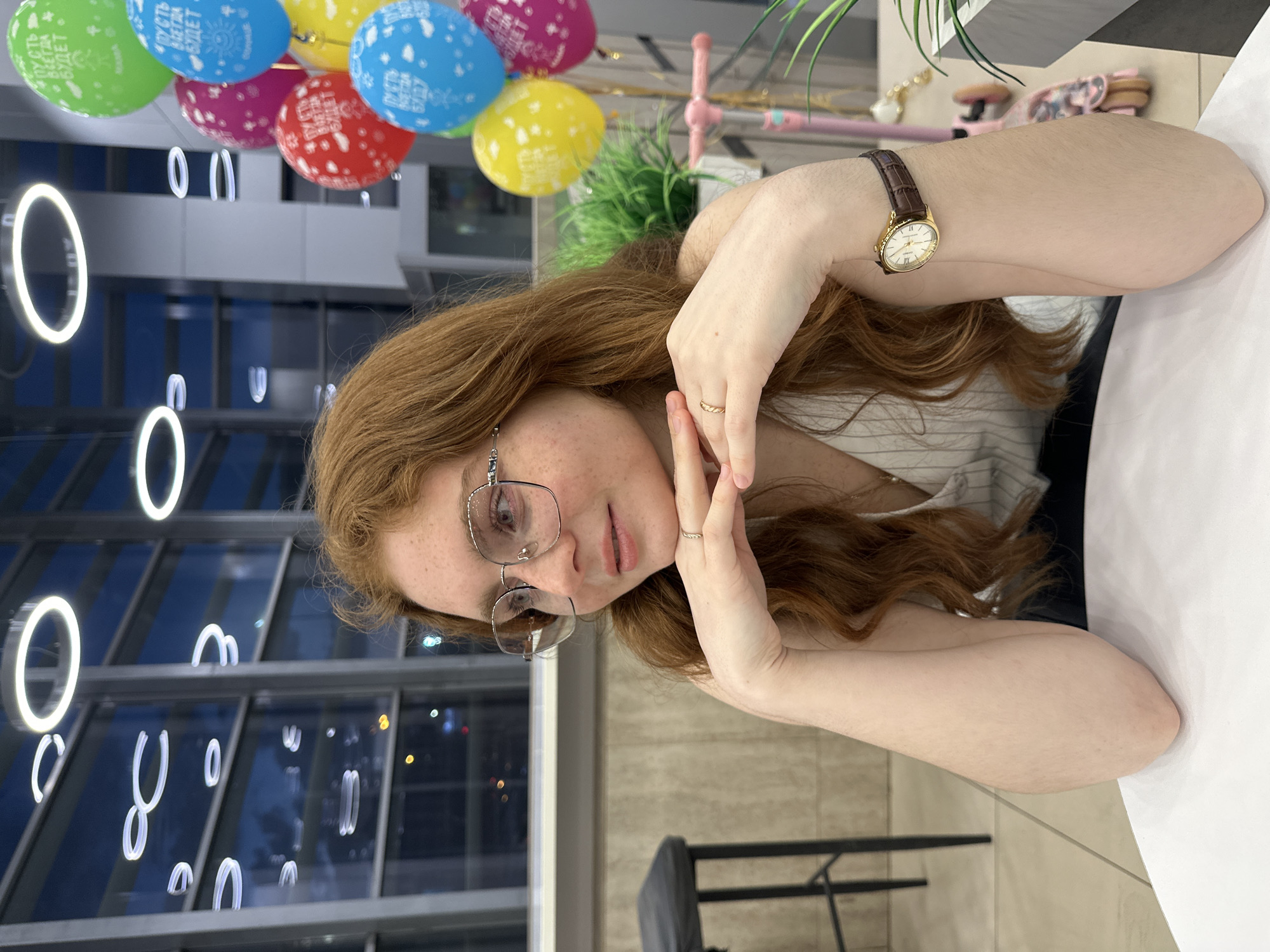 The image depicts a young woman with long, wavy reddish-brown hair and freckles, wearing glasses, as the central subject. She is looking towards the viewer with a calm expression, resting her head on her hands, which are clasped in front of her. On her left wrist, she wears a gold-colored wristwatch with a brown leather strap, and a simple gold ring adorns her right ring finger. She is dressed in a light-colored top with subtle vertical stripes.
The scene is an indoor setting, likely a modern cafe or restaurant in Kazan, Russia, characterized by a festive atmosphere. Numerous colorful balloons in green, blue, pink, yellow, and red are visible in the background. The text "ПУСТЬ ВСЕГДА БУДЕТ" (meaning "MAY IT ALWAYS BE") is clearly visible on some of the balloons, along with partial readings like "БУДЕТ" and "БАДУ," indicating a celebratory event.
The background features large reflective surfaces, possibly windows or decorative panels, showcasing abstract, glowing circular and U-shaped light fixtures. Beyond these, blurred lights in the distance suggest the time of day is evening or night. A hint of greenery, possibly a potted plant, and a small pink decorative object are also visible. A dark chair and part of a table are present in the foreground and background, adding to the indoor setting. The overall impression is a relaxed moment within a festive, contemporary environment.