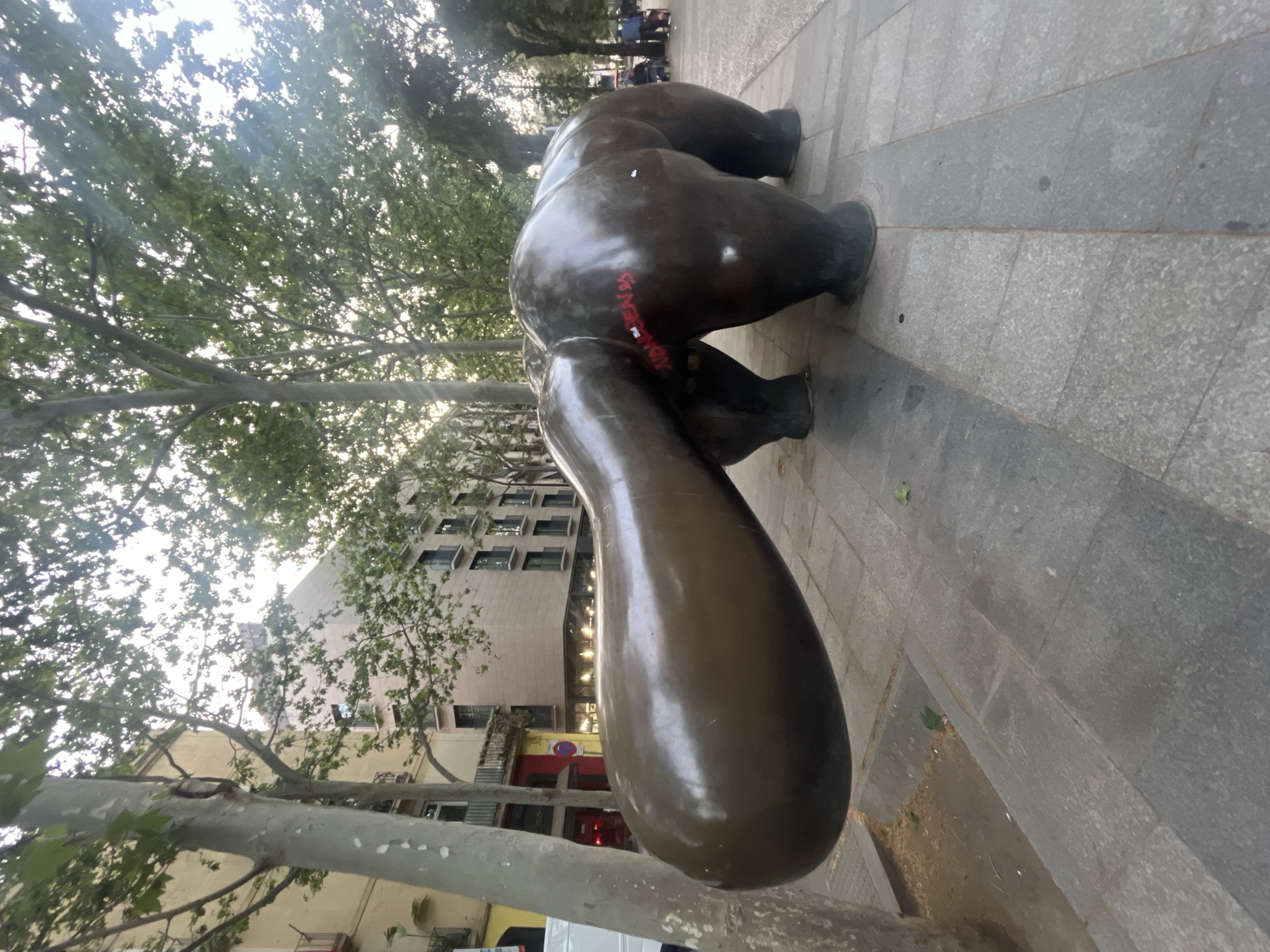 A bronze statue of a reclining woman is on a sidewalk in Barcelona, Spain. The statue is oriented with its rear end facing the viewer and features graffiti in red paint on the right buttock. The statue is positioned on a gray, stone-tiled sidewalk lined with trees and a building in the background. The photograph is taken from a low angle.