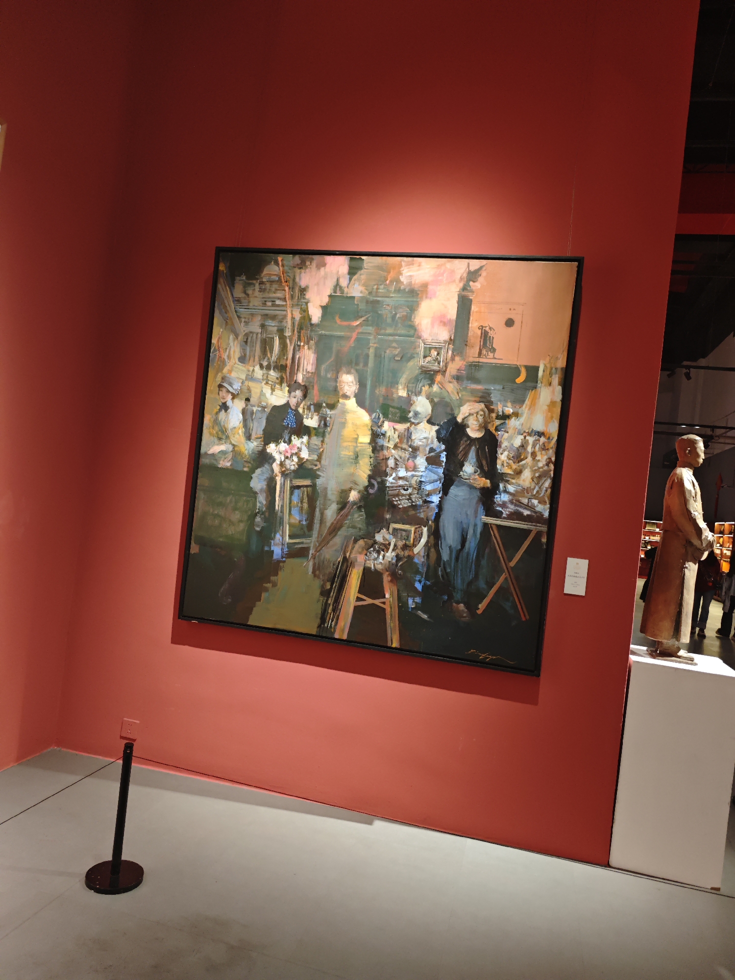 The image shows an art exhibition. The background features a red wall, on which hangs a framed painting depicting a group of figures in an indistinct, possibly historical or theatrical, setting rendered in a broad, impressionistic style. To the right of the painting is a sculpture of a man, appearing to be made of wood or bronze, positioned on a white plinth. In the foreground, a barrier post and rope indicate a boundary for the art display, and the floor is a neutral shade of grey. The gallery appears to be well-lit. The location seems to be inside a building in Unknown, Unknown.
