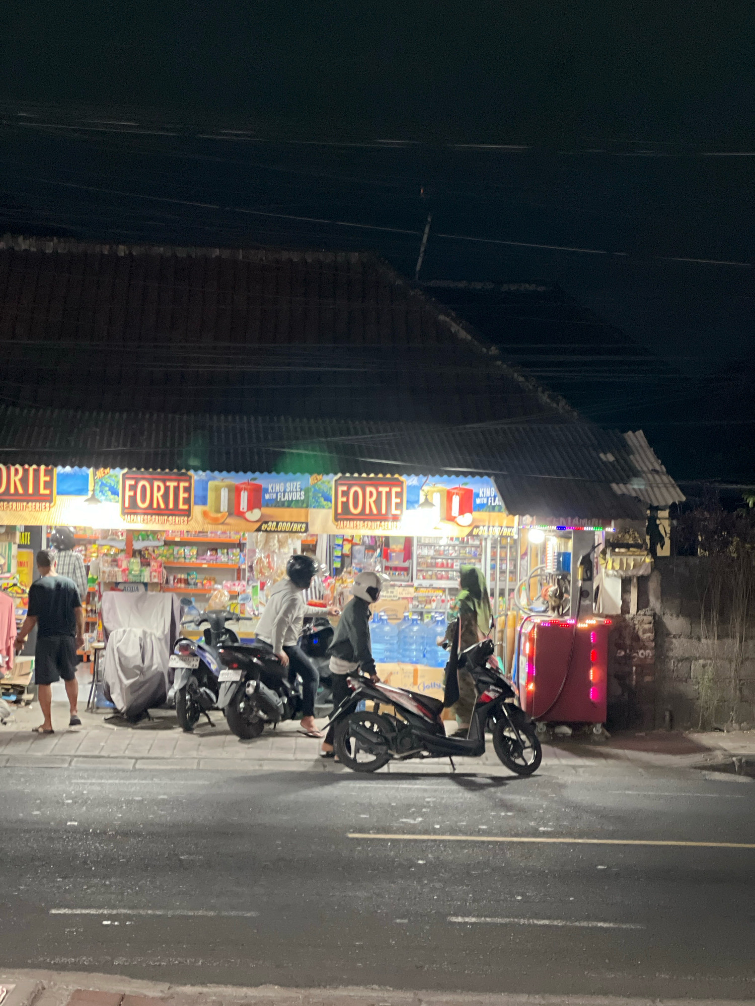 The image depicts a street scene at night in Denpasar, Indonesia, with a storefront advertising "FORTE" products, likely cigarettes given the "KING SIZE WITH FLAVORS" text visible. Several individuals are present, some standing and others on motorcycles. Two motorcycles are parked in front of the store, and a third is partially visible to the right. The storefront is illuminated, revealing shelves stocked with various items, including bottled water. A red, illuminated cooler or display unit is positioned to the right of the storefront. The street in front of the store is paved, with a yellow line marking the lanes. Overhead, power lines are visible against the dark night sky. The overall environment suggests a commercial area that remains active after dark.