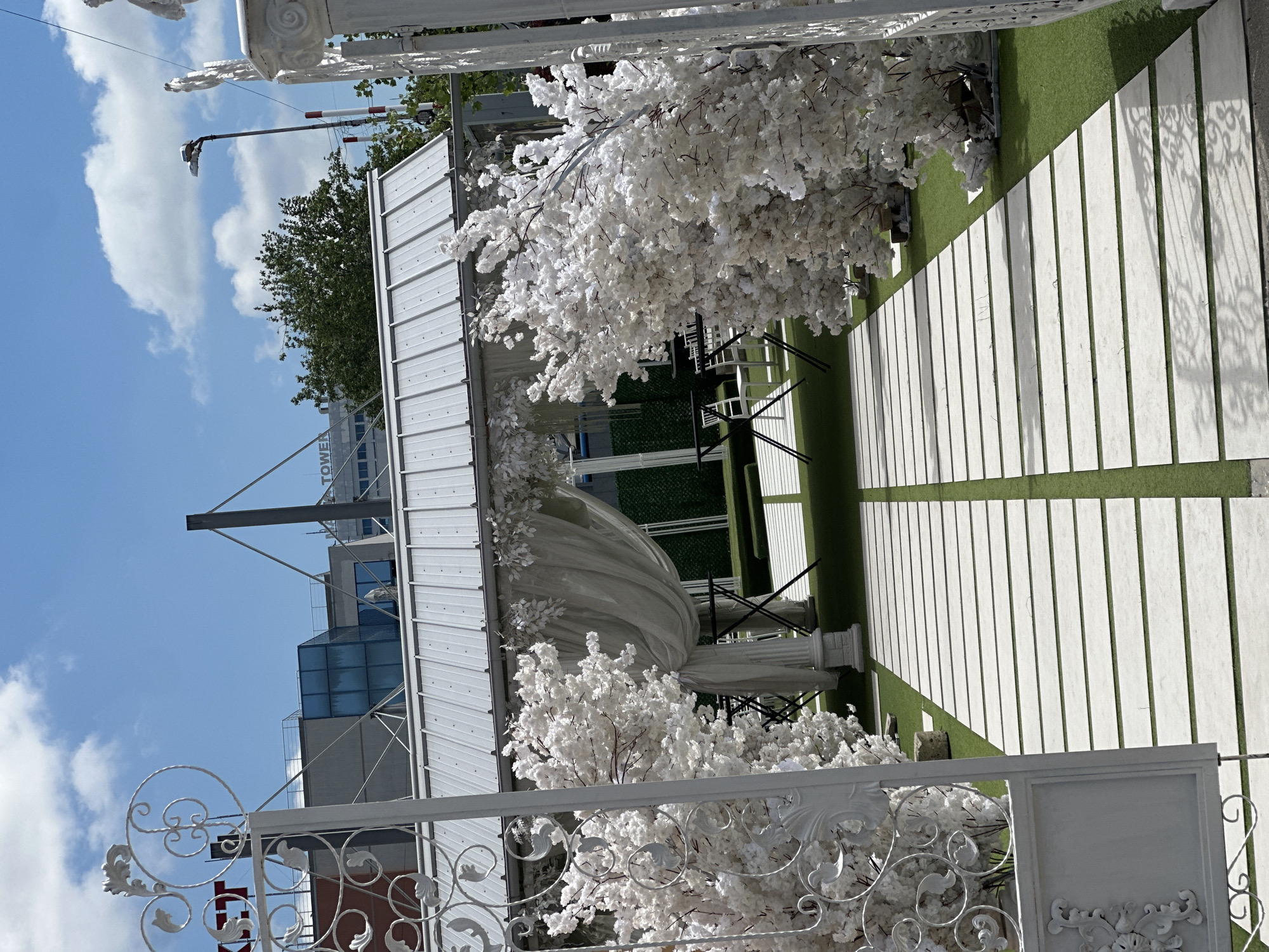 A bright, outdoor setting displays a decorated wedding area. A white metal gate with ornate scrollwork opens to a walkway of rectangular white slabs bordered by green artificial turf. On both sides of the path, artificial white blossom trees stand. A white gazebo, draped with white fabric, sits over black chairs. The background shows buildings, including one labeled "TOWER." The location is Pleven, Bulgaria.