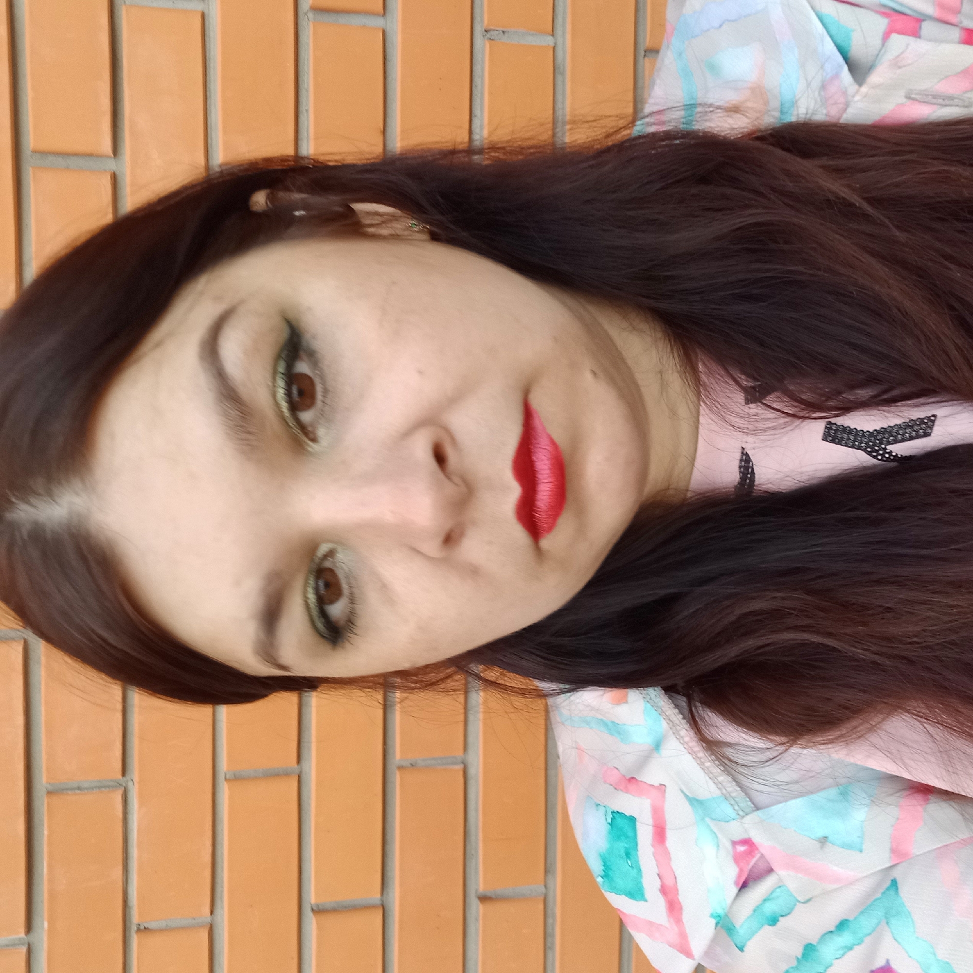A medium shot captures a woman's face from the chest up, positioned centrally against a brick wall. She has dark brown hair, some of which frames her face while the rest cascades down her shoulders. Her eyes are brown and adorned with green eyeshadow and black eyeliner. Her lips are painted a vibrant red. She is wearing a light-colored jacket with a geometric pattern of pink, blue, and white, over a pink shirt. The background consists of an orange brick wall. The image appears to be taken outdoors, possibly in Kropyvnytskyi, Ukraine, given the typical architecture.