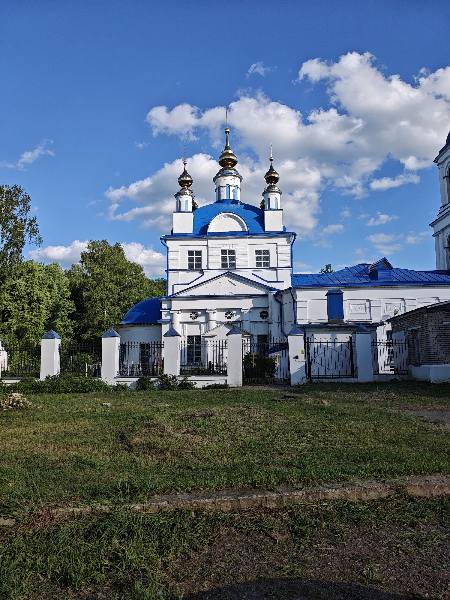 The image depicts a Russian Orthodox church with white walls and blue roofs and domes. The church has several golden spires. A black wrought-iron fence surrounds the church property, with grass in the foreground. Trees are visible behind the church, and a blue sky with fluffy white clouds provides the backdrop. The image was taken in Мамонтово, Russia.