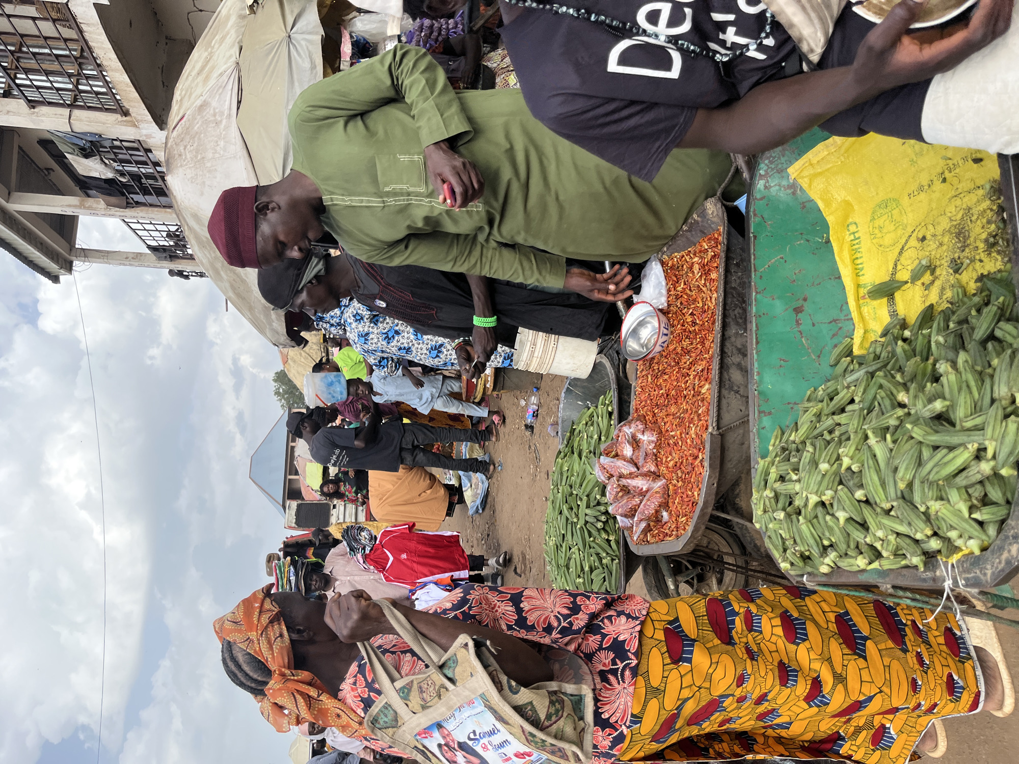 This media file depicts a bustling outdoor market scene in Rafin Par, Nigeria. The image is oriented vertically, presenting a dynamic view of people and goods.
Several individuals are visible, some interacting with each other and others engaged in managing the market stalls. One person in the foreground wears a distinctive orange headscarf and a colorful patterned dress, carrying a tote bag adorned with text and an image. Another individual to their right wears a yellow and red patterned wrap dress. In the center of the frame, a man in a green traditional outfit and a maroon cap is seen alongside others dressed in various casual attire, including a black t-shirt with white text. The presence of multiple individuals suggests a public gathering or a busy shopping day.
Objects in the scene include a variety of produce displayed on a cart. A large pile of fresh green okra is prominent, alongside containers filled with dried red chilies. A yellow plastic bag, possibly used for packaging or display, rests near the okra. A metal bowl sits near the chilies. The market appears to be set up along a dirt path, with stalls and buildings suggesting a well-established marketplace.
The sky is visible in the background, showing scattered clouds, indicating it's daytime, likely in the morning or afternoon. The overall atmosphere appears lively and transactional, typical of a local market. The vibrant colors of the clothing and produce contribute to the energetic feel of the scene.
Visible text on the tote bag appears to be in English and includes the names "Samuel & Jamm" with a wedding-like image. The yellow plastic bag has some text, possibly indicating a brand or origin, with "CHIKUN" being partially discernible. The black t-shirt has white text that reads "Dè."
