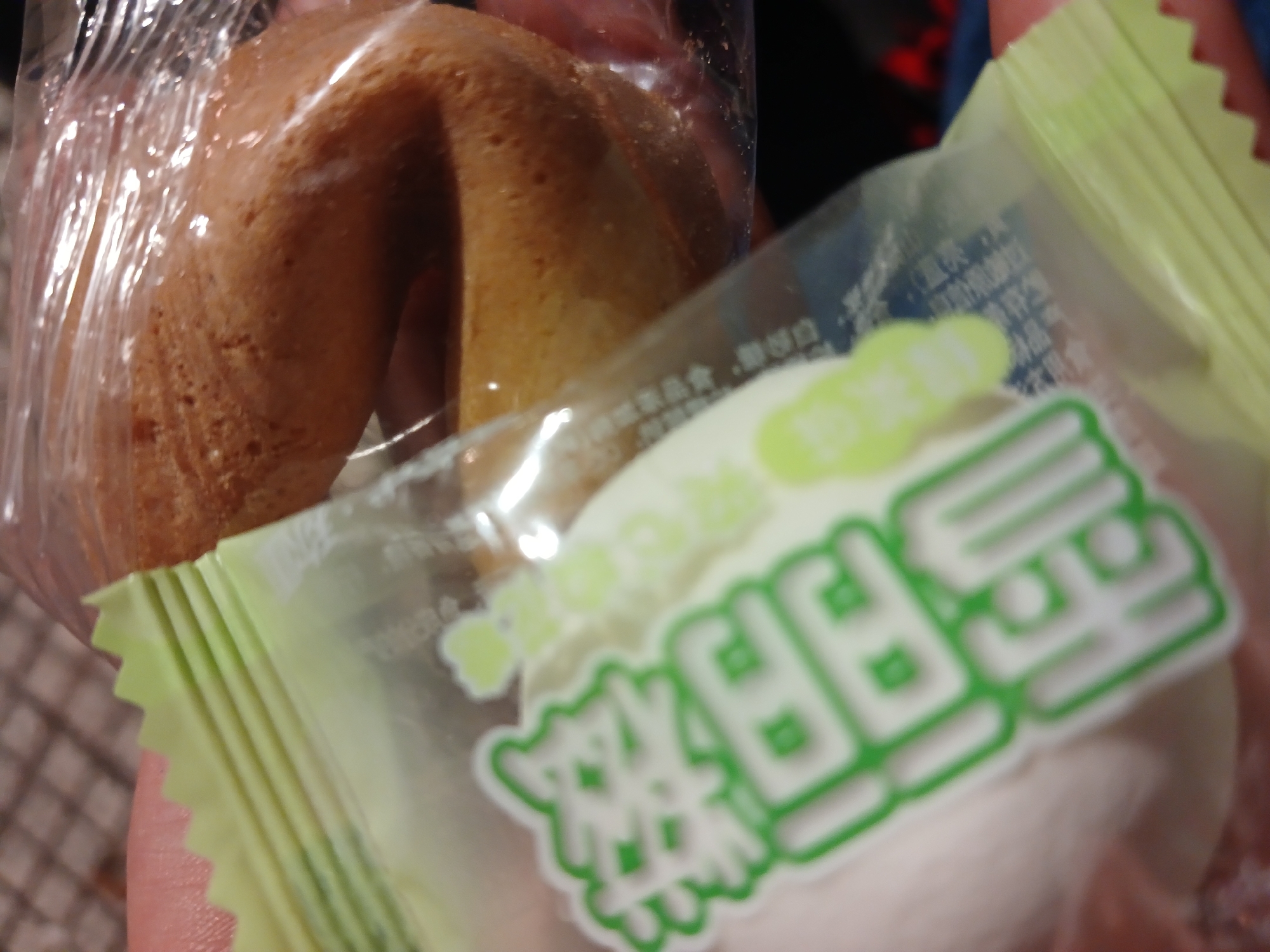 The image shows a hand holding two individually wrapped treats. One is a fortune cookie, encased in clear plastic. The other appears to be a marshmallow or similar confection, sealed in a pale green package with Asian characters printed on it. The background is blurred, but reveals a hint of a tiled surface and an area with unclear colors. There is no clear indication of Buenos Aires or any other location.