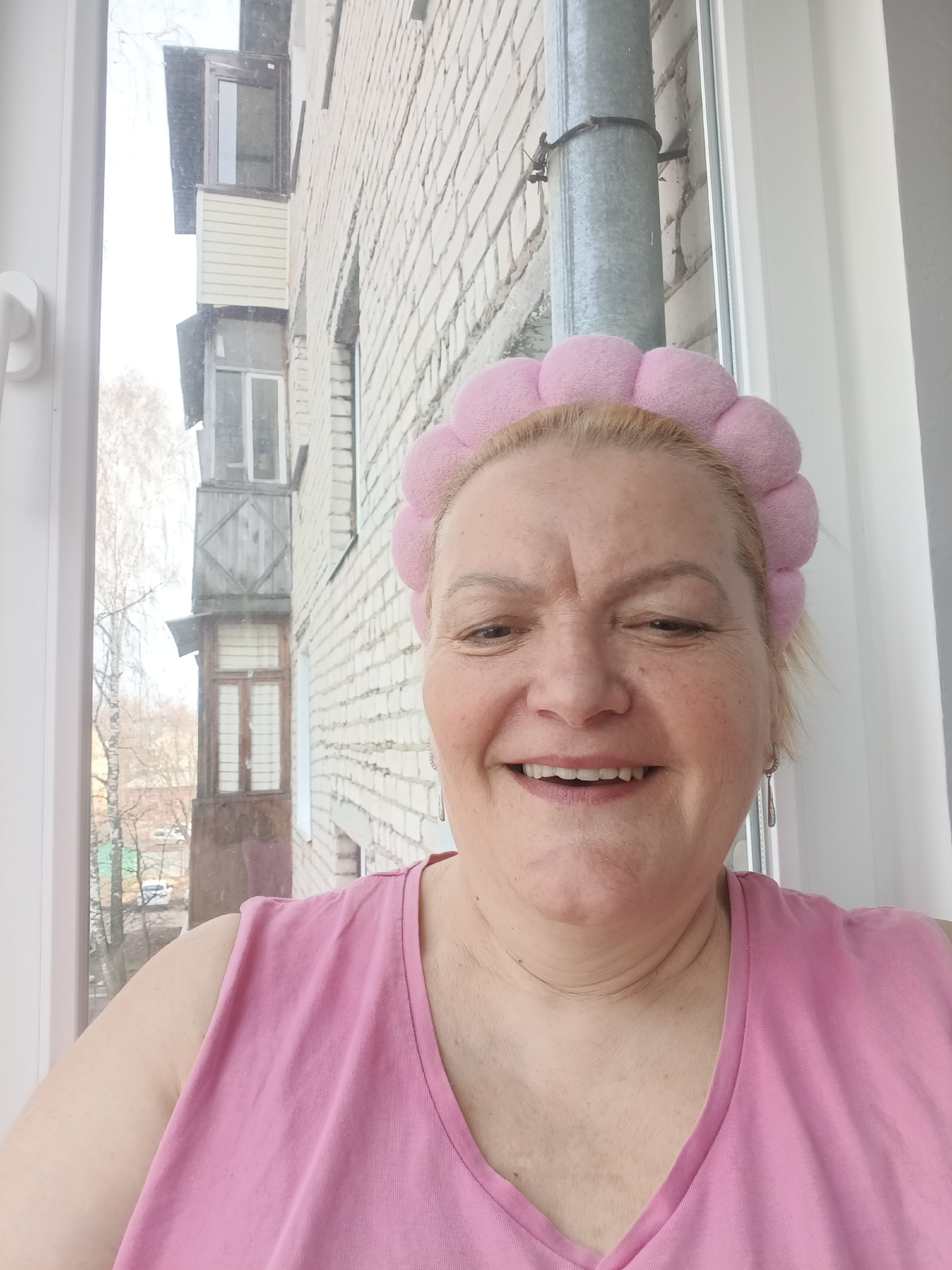 A middle-aged woman with blonde hair and a pink headband smiles at the camera while standing inside a room with a window. She is wearing a pink tank top. Outside the window is a view of a light brick building, a metal pipe, and a tree without leaves. Further in the background, there is another building with wooden balconies. A street with cars is visible between the buildings. The location is Murom, Russia.