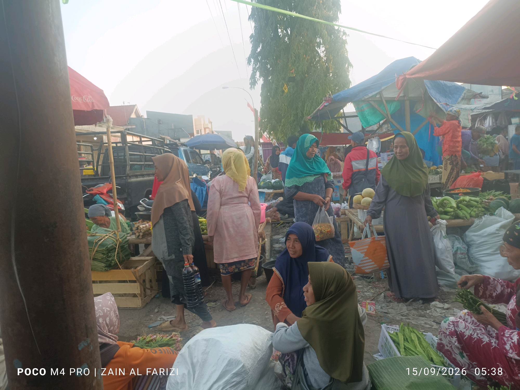 An outdoor marketplace is depicted in Arosbaya, Indonesia. Several individuals, predominantly women wearing hijabs, are present, engaged in various activities. Some are standing and conversing, while others are seated, interacting with merchandise. The marketplace appears to be a produce market, with an abundance of fruits and vegetables, including green beans, watermelons, and what appear to be bundles of leafy greens. Several makeshift stalls or tables are visible, covered with tarps and fabrics to provide shade. In the background, trucks and a red Coca-Cola umbrella suggest the presence of goods being transported or sold. The ground is a dirt and pavement mix. The overall atmosphere suggests a bustling day at the local market.