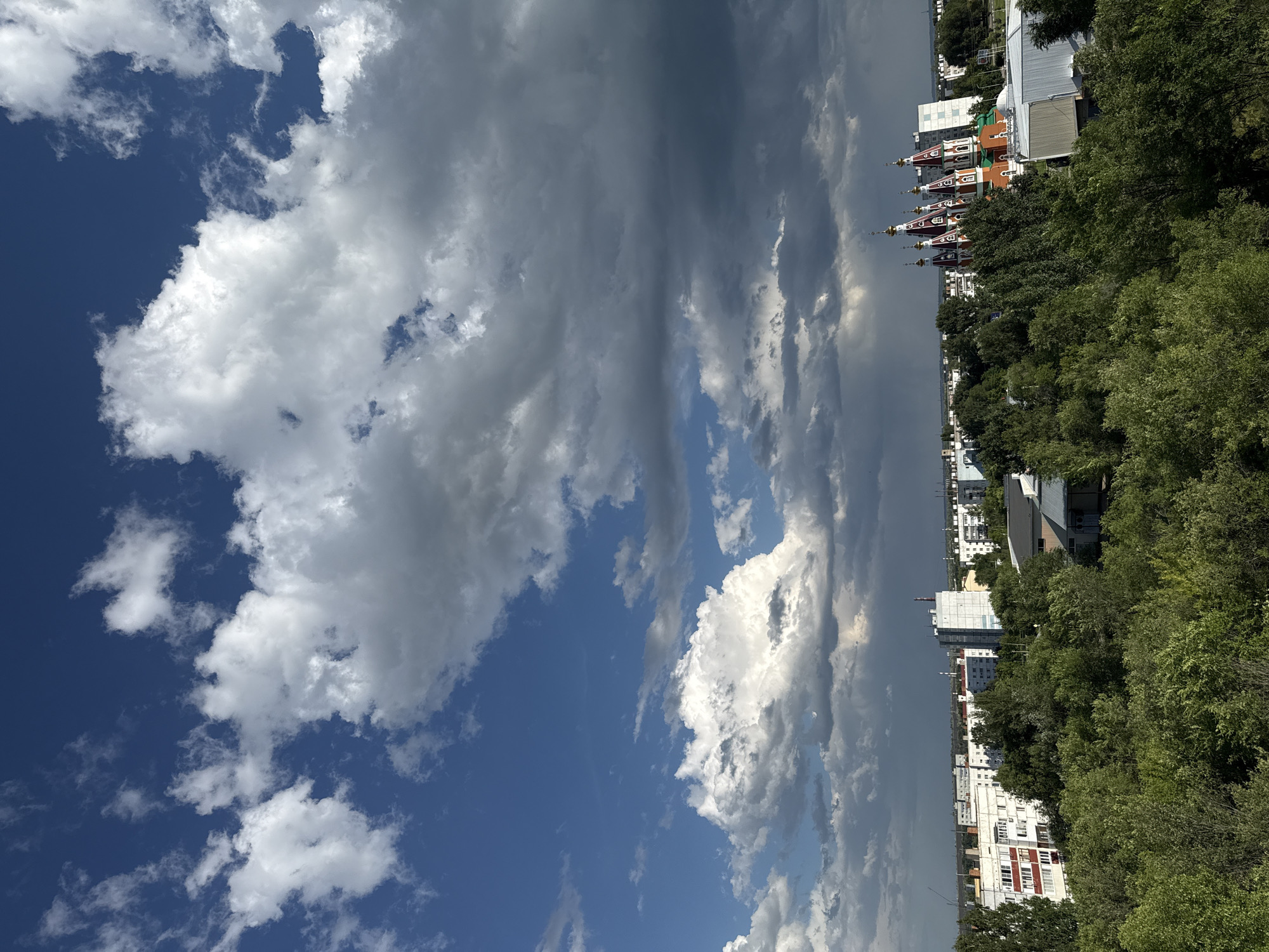 The image shows a view of the city of Otradny, Russia, with a dramatic sky filled with white and grey clouds. In the foreground, a dense canopy of green trees fills the lower half of the frame. Behind the trees, a cluster of multi-story residential buildings and some smaller structures are visible. Dominating the right side of the mid-ground is a colorful church with multiple golden domes and red and white accents. The sky above transitions from a deep blue on the left to darker, heavier clouds on the right, suggesting an approaching storm. The overall scene depicts an urban landscape partially obscured by lush vegetation under a dynamic sky.
