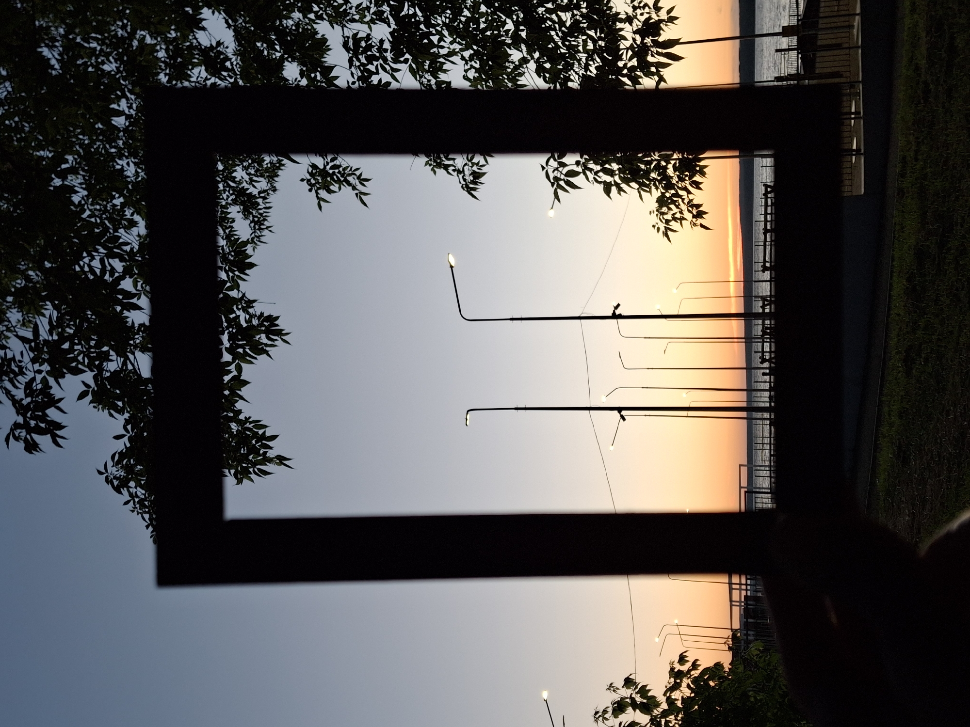 A silhouette of a rectangular frame held by a person's hand is in the foreground, obscuring a portion of the view. Through the frame, a twilight sky transitions from a pale blue at the top to a warm orange glow near the horizon. Several streetlights are visible, their lamps illuminated. A body of water, likely the Kama River, stretches across the middle ground, with a distant shoreline. A pier or embankment with benches and railings is situated along the water's edge. Trees with dark leaves frame the top and sides of the image, their silhouettes stark against the fading light. The scene is set in the city of Chaikovsky, Russia.