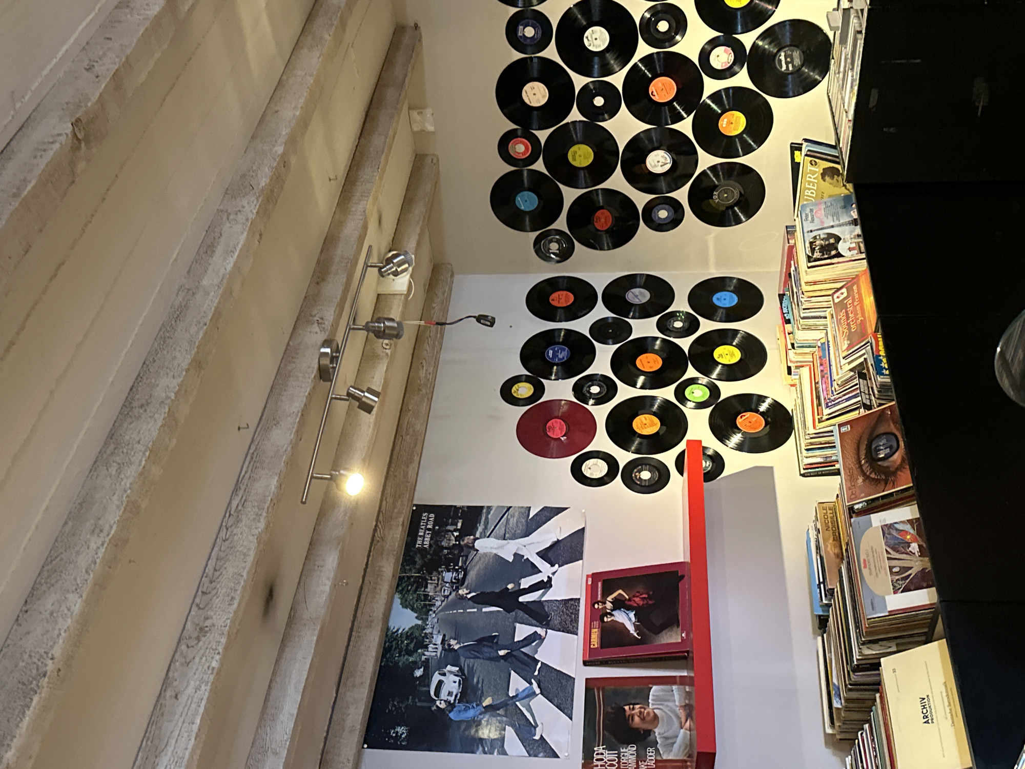 The image depicts the interior of a record store. The walls are adorned with a collection of vinyl records, some of which are displayed as decorative items, while others are visible in the record bins at the bottom of the frame. A large poster of The Beatles' Abbey Road album cover hangs on the wall, and two album covers are displayed on a red shelf below it. The ceiling features exposed wooden beams and track lighting. The record bins are filled with numerous record albums, showcasing various cover art. The overall impression is that of a well-stocked and visually interesting music shop. The scene is set in Bruges, Belgium.