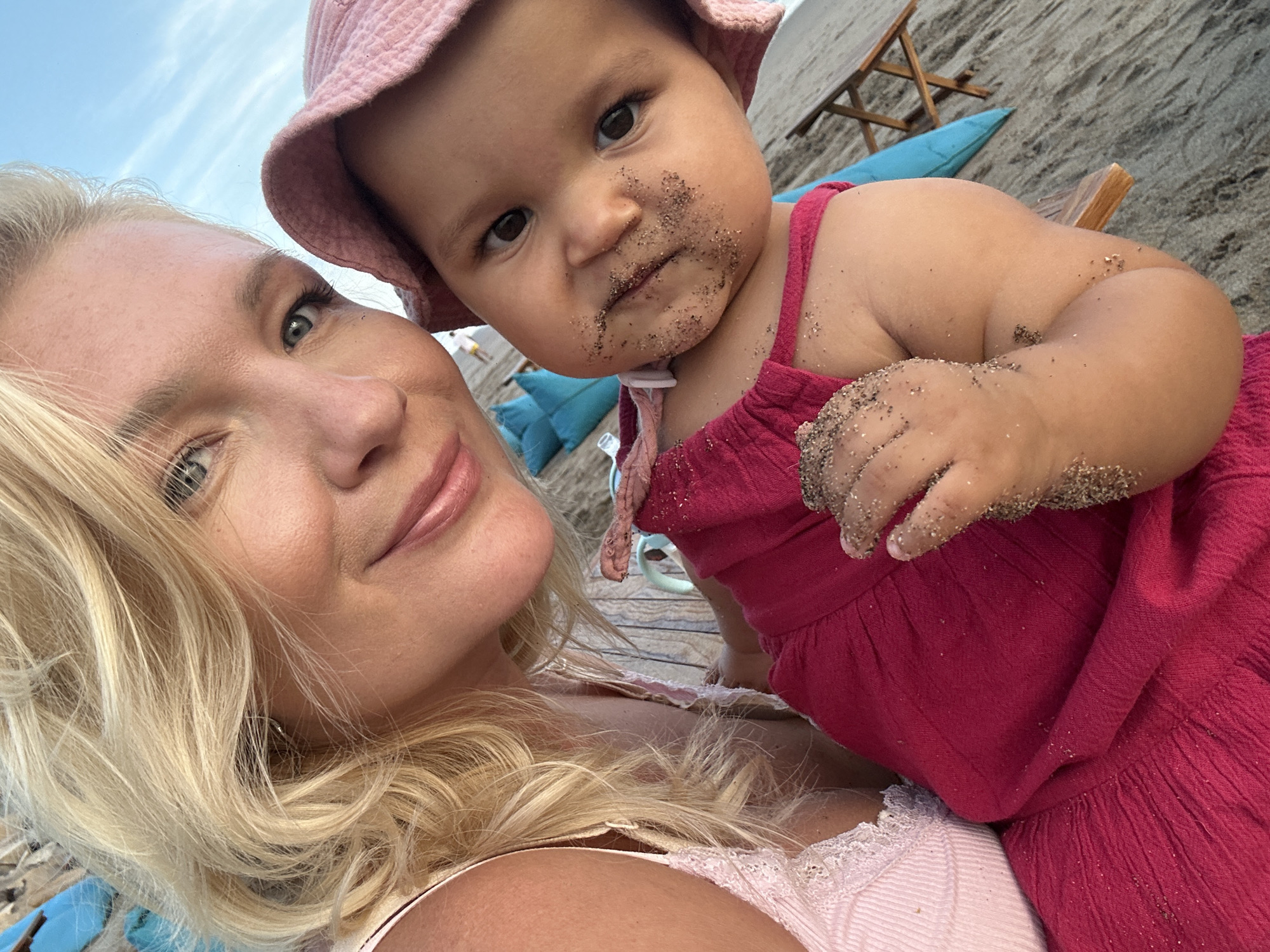 A blonde woman takes a close-up selfie with an infant on a beach in North Kuta, Indonesia. The woman is fair-skinned with blonde hair, wearing a light pink top with lace trim. The infant, wearing a pink sun hat and pink dress, has sand around their mouth and on their hand. The background shows a wooden deck, sand, beach chairs, and blue pillows. The sky is partially visible in the upper left corner.
