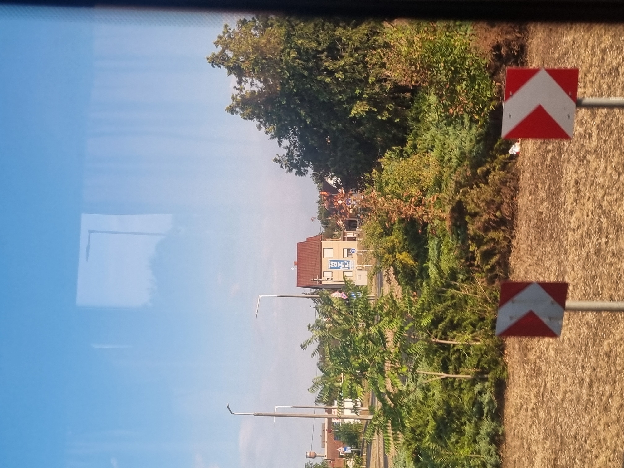 The image depicts a roadside scene in Berettyóújfalu, Hungary, likely on the outskirts of the city. In the foreground, a brown, dry, grassy field is visible, with two red and white directional signs pointing to the right. Behind the field, a dense area of green and autumnal foliage forms a natural barrier. Beyond the vegetation, several buildings are present, including a multi-story building with a red roof that appears to be a hotel, indicated by a blue "HOTEL" sign. Other structures, possibly houses or smaller commercial buildings, are also visible, along with streetlights and some traffic signs. The sky is clear and blue, suggesting daytime. The overall impression is one of a suburban or rural roadside environment.