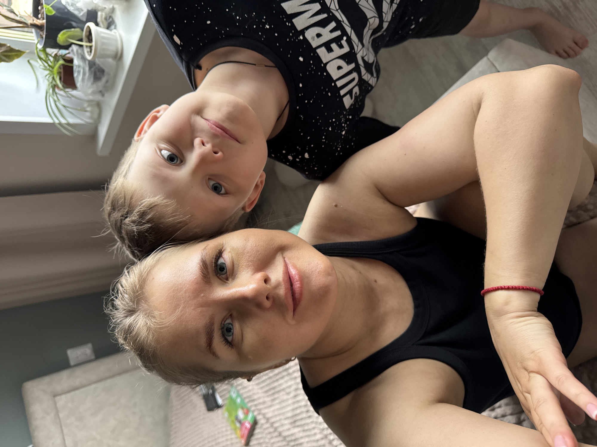 A woman and a child are posing close together for a selfie-style photograph taken indoors. The woman is in the foreground wearing a black tank top and a red bracelet. The child is behind her, wearing a black shirt with white writing. The background includes a window with plants on the windowsill and a partial view of a room with a neutral color scheme. The location is Ферма, Russia.