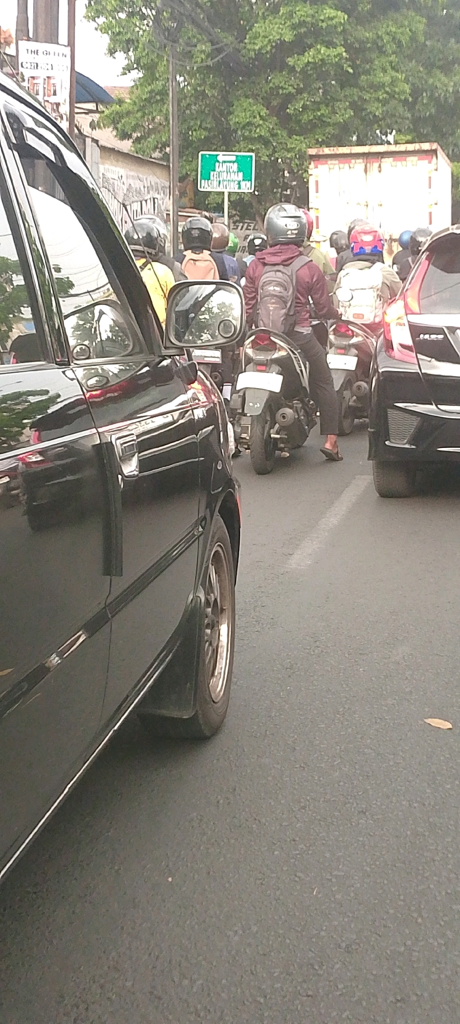 The image captures a street scene in Bandung City, Indonesia, characterized by moderate traffic congestion. In the foreground, the left side of a black car, likely a sedan, is prominently featured. Its reflective surface shows a blurred reflection of the surroundings. The car's wheel and fender are visible, with a mudguard attached.
The road is asphalt, with a white line indicating a lane divider. Several motorcycles are present, with riders wearing helmets. One motorcycle in the center of the frame has a rider wearing a maroon jacket and a backpack. Another motorcycle behind it has a rider in a yellow shirt. To the right, the rear of a black car, possibly a hatchback, is visible.
Above the traffic, a green street sign is mounted on a pole. The sign is in Indonesian and reads "KANTOR KELURAHAN PASIRKAMPING KM." This indicates the presence of the Pasirkampung Village Office nearby. Trees with dense green foliage line the street, creating a shaded environment. In the background, a large white truck with a patterned side is parked or moving slowly. The overall impression is a typical urban street in Bandung during daylight hours, with the characteristic blend of vehicles and signage.