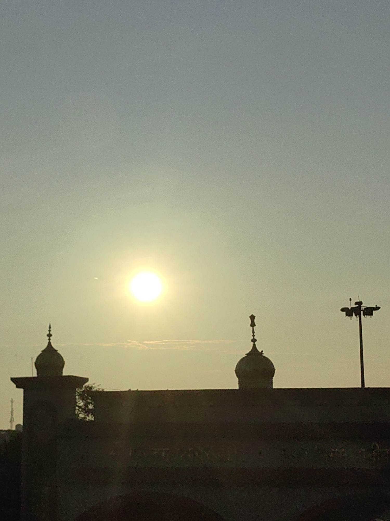 The image captures a silhouetted view of a structure, likely historical, against a hazy, pale blue sky. The sun is prominently visible, a bright, luminous orb in the upper left quadrant, casting a soft glow and creating a slight lens flare. The silhouette reveals architectural features characteristic of Islamic or Mughal design, specifically two onion-shaped domes topped with spires. These domes rise from a flat, dark structure that spans the lower portion of the image. To the right, a tall pole with multiple lamps is also silhouetted against the sky. The scene appears to be set in Panipat, India, a city known for its historical significance and architecture. The overall atmosphere is serene and contemplative, hinting at either dawn or dusk.