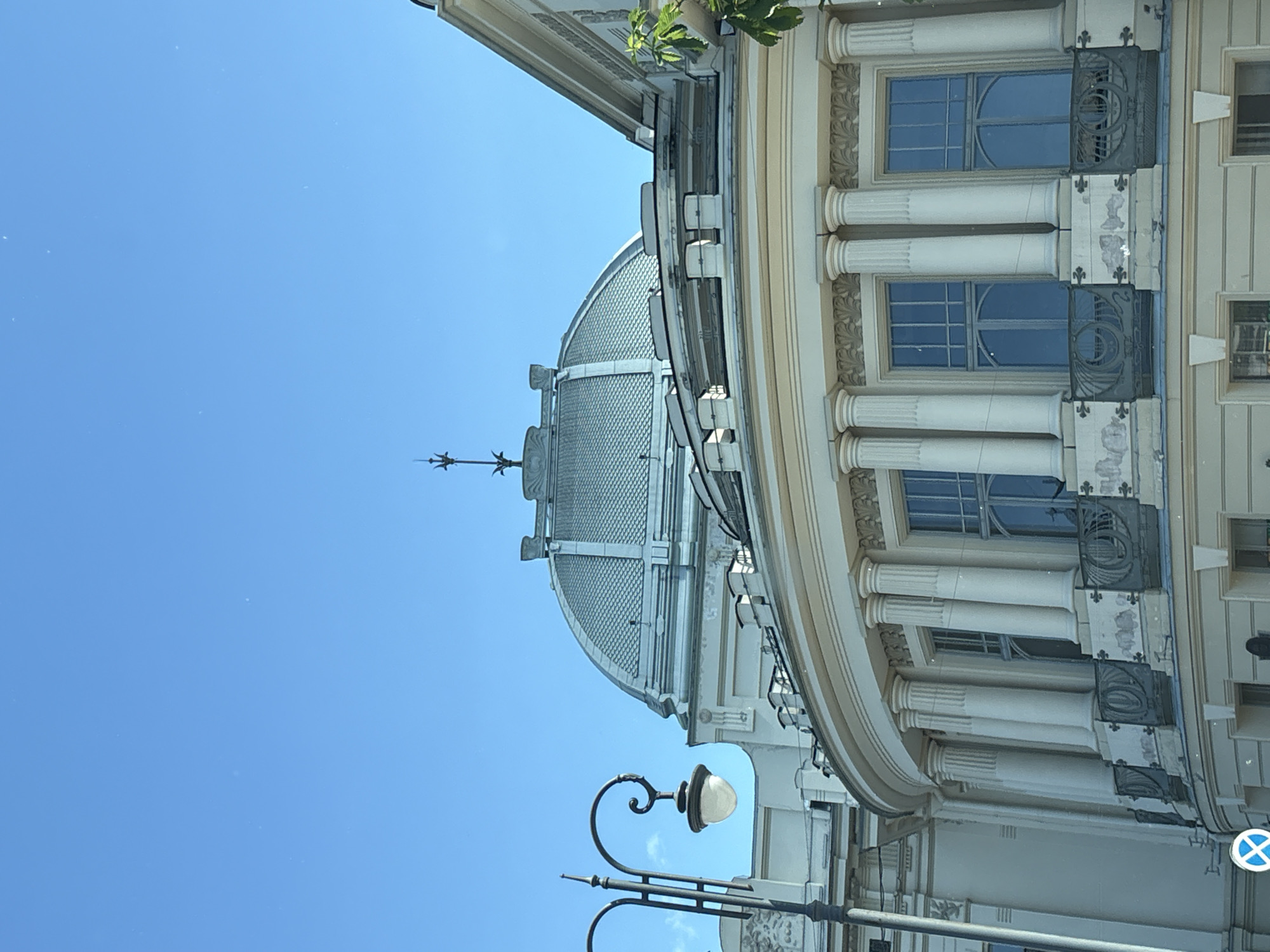 The image is a close-up shot of a domed building in Saint Petersburg, Russia. The building appears to be made of light-colored stone and features classical architectural elements such as columns and ornate balconies. A prominent dome with a patterned roof is visible, topped by a spire. A street lamp with a decorative curved arm and a glass lantern is positioned to the left of the frame, partially obscuring the building's facade. The sky is clear and blue. The lower part of the building shows several large windows with decorative iron railings on small balconies.