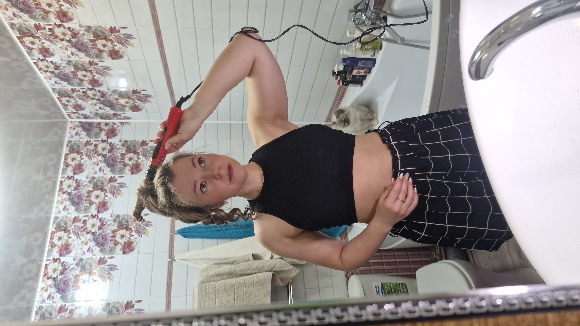 A young woman is captured in a mirror selfie, styling her hair with a curling iron in what appears to be a home bathroom. The image is oriented with the woman's head to the right and her feet to the left, as if she is looking up into the mirror.
The woman has light-colored hair, some of which is already styled into bouncy curls, and wears a black sleeveless crop top revealing her midriff, paired with black shorts featuring a white grid pattern. She holds a red and black curling iron in her right hand, using it on a section of her hair near her temple. Her expression is neutral and focused on her task. A simple gold-colored band adorns her left ring finger, and her fingernails have a light-colored polish.
The bathroom features white wall tiles, with the upper portion decorated with a vibrant floral pattern in shades of purple, brown, and cream. The lower walls are a plain light grey. In the background, a white bathtub is visible with a chrome faucet and showerhead. Several bottles of bath products are lined up on the edge of the tub. A fluffy, light-colored cat sits calmly on the tub's rim, observing the scene. A white toilet is partially visible behind the woman, to the right. A blue towel and a cream-colored towel hang on a rack. A white bottle with the visible text "nahsa" rests on the floor. The reflection of the floor shows a reddish-brown checkered pattern.
The scene depicts a casual, personal moment of grooming, likely in Otchor, Russia, given the provided location context.