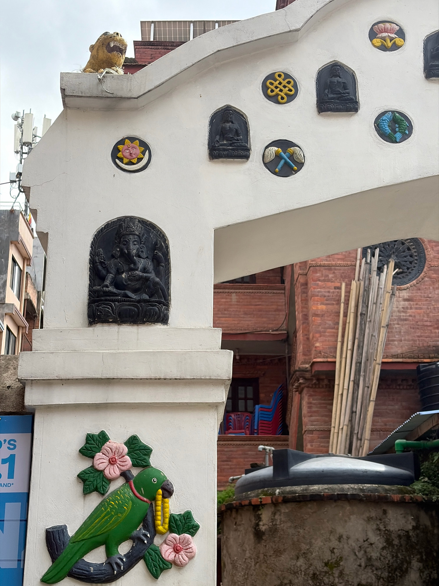 This image depicts an ornate archway decorated with various carvings and sculptures, likely in the city of Lalitpur, Nepal. At the top of the arch, a lion or tiger sculpture with an open mouth is visible. The archway itself is white and features several circular decorative elements. These include a sun and moon motif with faces, a Buddhist endless knot, a seated Buddha figure, a pair of crossed symbolic objects, a pink lotus flower, and a pair of blue and green fish. Below, on a white pillar, a carved relief of the Hindu deity Ganesha is prominently displayed. Further down, on the left side of the pillar, a colorful sculpture of a green parrot perched on a branch is depicted, holding a yellow beaded necklace in its beak, surrounded by pink flowers and green leaves. In the background, a red brick building with windows and balconies can be seen, along with stacked colorful chairs. A collection of bamboo poles and a water tank are also visible. The overall scene suggests a location with cultural and religious significance, common in the Kathmandu Valley.