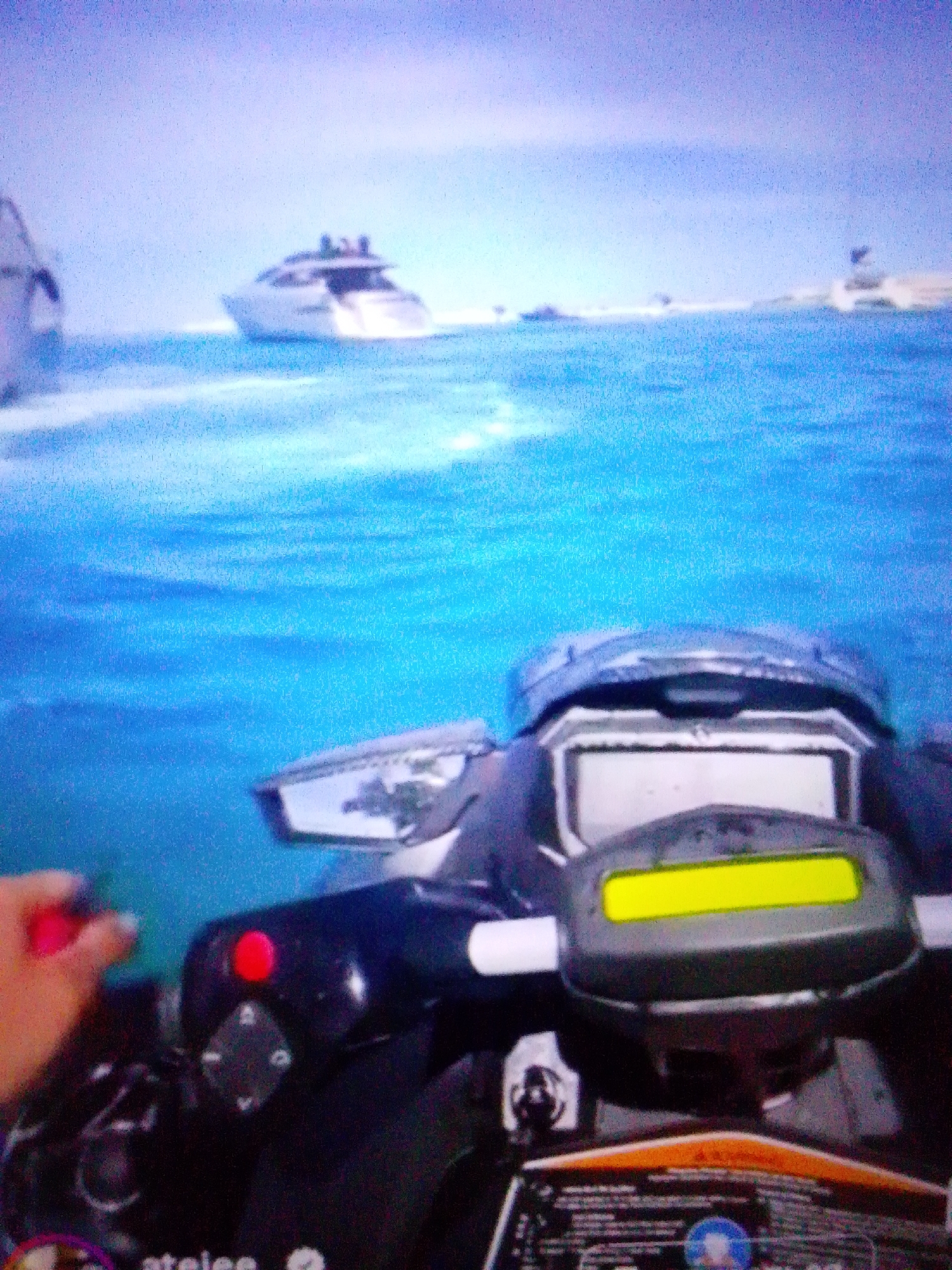 The image shows a first-person perspective of someone riding a jet ski on a body of water. In the foreground, the handlebars and dashboard of the jet ski are visible, including a red button and a yellow display screen. A hand is seen reaching for the red button. In the background, a large white yacht with several people on deck is sailing on the water. The sky is a pale blue with some clouds. The location is not Aznakayevo, Russia.