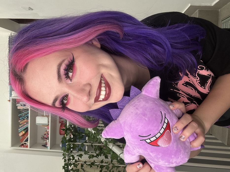 An indoor portrait shows a smiling female in the City of Zagreb, Croatia, looking directly at the camera. Her long, wavy hair is dyed vibrant pink on the top and front sections, transitioning to dark purple for the remainder. Her eyes feature pink eyeshadow, prominent dark winged eyeliner, and false eyelashes, while her lips are colored with dark red lipstick. She wears a black t-shirt adorned with a light-colored graphic print. She holds a light purple plush toy, resembling a stylized Pokémon character with an open-mouthed smile, red tongue, and white teeth, in both hands. Her fingernails display a light-colored polish with small decorative elements. The background on the left features a white multi-shelf bookcase stocked with books and various small decorative objects, including a rectangular mirror. An artificial green ivy vine is draped vertically to the left. The right side of the background is a plain white wall, with a dark mat and a light-colored step partially visible at the bottom right.