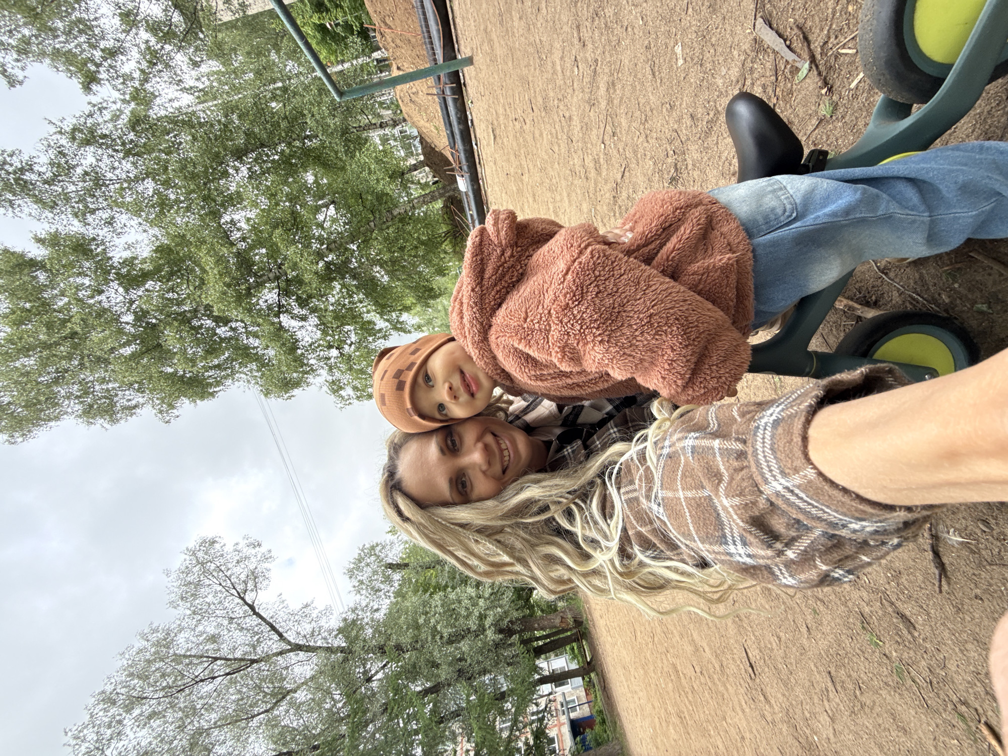 A woman with long blonde hair is taking a selfie with a toddler in an outdoor setting. The woman is wearing a brown and white plaid shirt, and the toddler is wearing a brown hat and jacket. They appear to be on or near a green tricycle. The ground is dirt or sand, and trees are visible in the background. The location is potentially in Rusanovo, Russia.