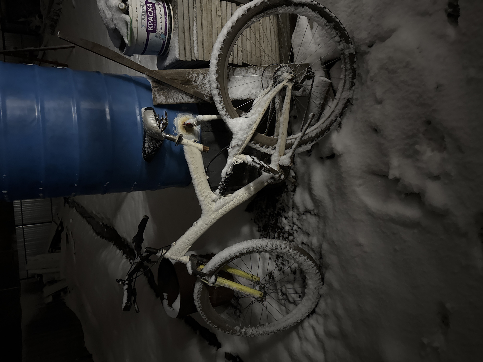 The image shows a snow-covered bicycle leaning against a blue barrel, with a bucket labeled "KPAСKA" resting on top of wooden planks nearby. The scene appears to be outdoors, likely in the city of Chaikovsky, Russia. The ground is blanketed in snow, and the bicycle is also covered in snow, suggesting a recent snowfall. Other dark objects are visible in the background.