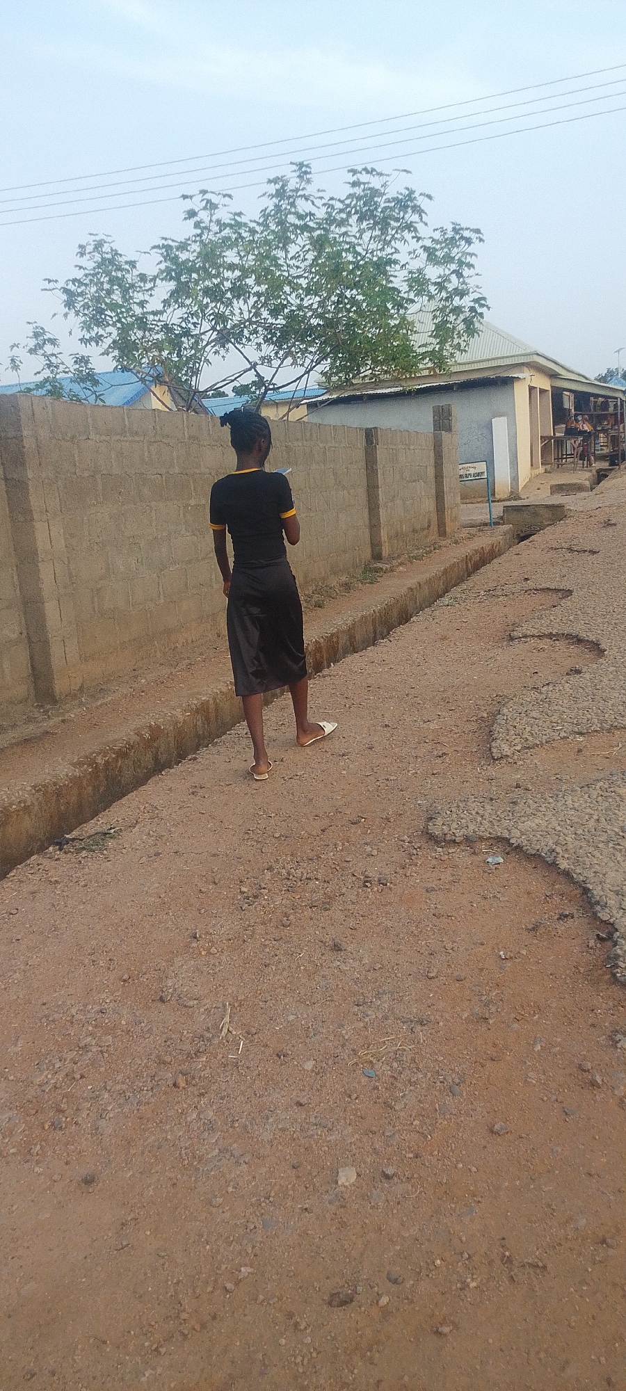 A person stands on a dirt road in Adankolo, Nigeria. The individual wears a black top with yellow trim, a long black skirt, and white sandals. They face away from the viewer. To the left is a concrete block wall. The road shows signs of wear and tear with patchy asphalt sections. In the background is a building with a silver roof and a sign, partially obscured, with "DIVINE FAITH ACADEMY" visible. Above the wall and building are trees and a light sky.