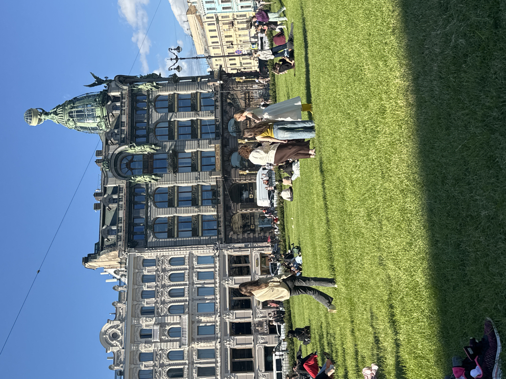 This is an eye-level, medium shot of Nevsky Prospekt in Saint Petersburg, Russia, on a sunny day with a clear blue sky and some clouds. The foreground is dominated by a vast, well-maintained green lawn, where several groups of people are casually sitting, lying down, and standing. In the mid-ground, a diverse group of individuals, predominantly women, are gathered in a huddle, engaged in conversation. One woman in a light-colored top and dark pants walks away from the viewer towards the right.
Behind the lawn, a row of ornate, historic buildings forms the backdrop. The most prominent building, on the right side of the frame, is the Singer House (Dom Knigi), recognizable by its striking green, dome-shaped rooftop topped with a glass globe supported by an eagle sculpture. This building features intricate architectural details, including numerous windows and decorative elements. To its left, another grand building with a white facade and arched windows stands tall. Beyond these, other buildings of varying architectural styles and colors line the street.
The scene depicts a lively urban environment where people are enjoying a pleasant day outdoors amidst the city's rich architecture. The presence of people spread across the lawn suggests a public space being utilized for relaxation and social interaction. Traffic is visible in the background, indicating that this is a bustling city thoroughfare.